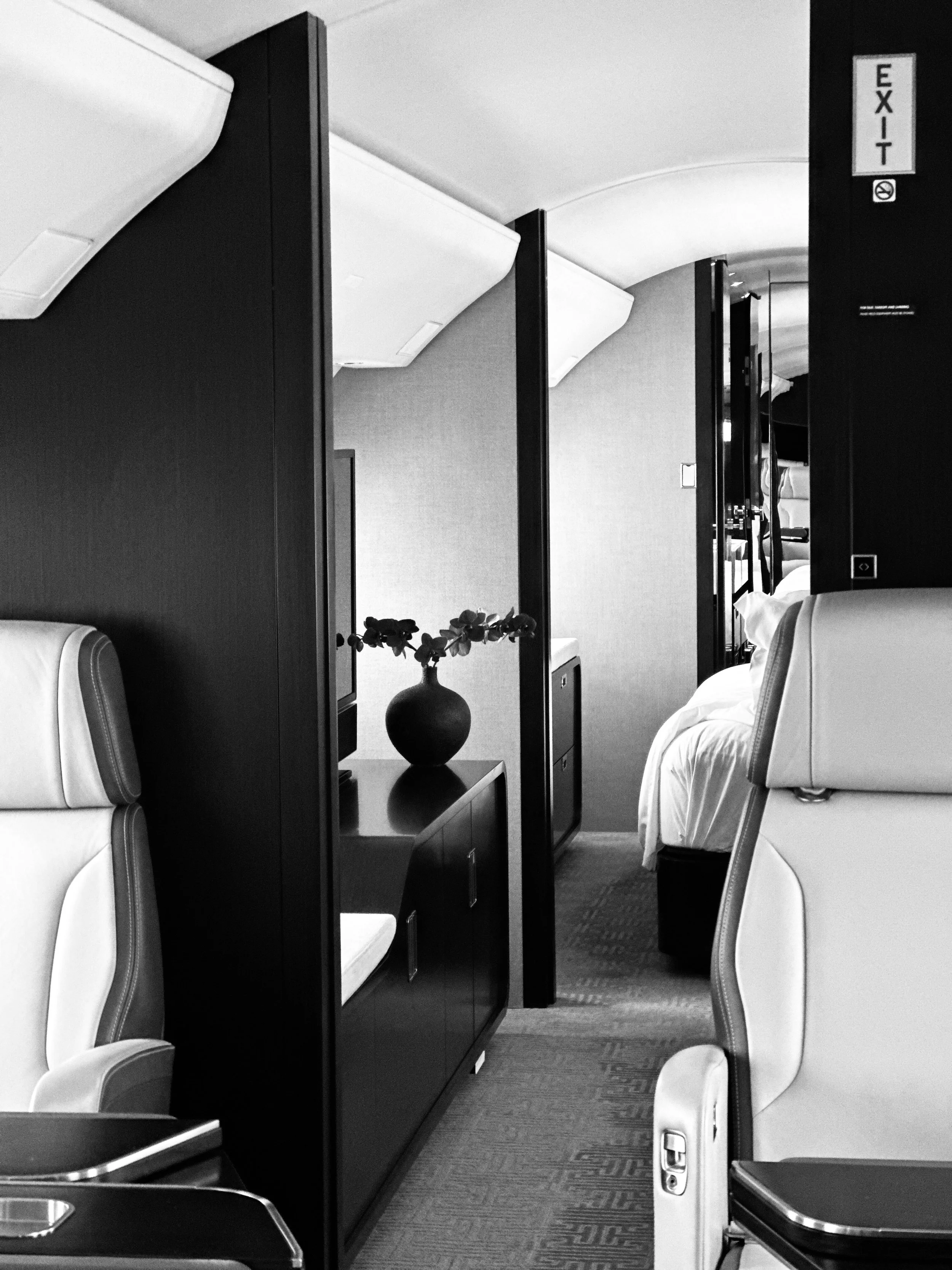 Inside an airplane cabin, showing a seating area with a small table, a TV, and a vase with flowers, and a view into a bedroom with a bed and side tables. Black and white photo.