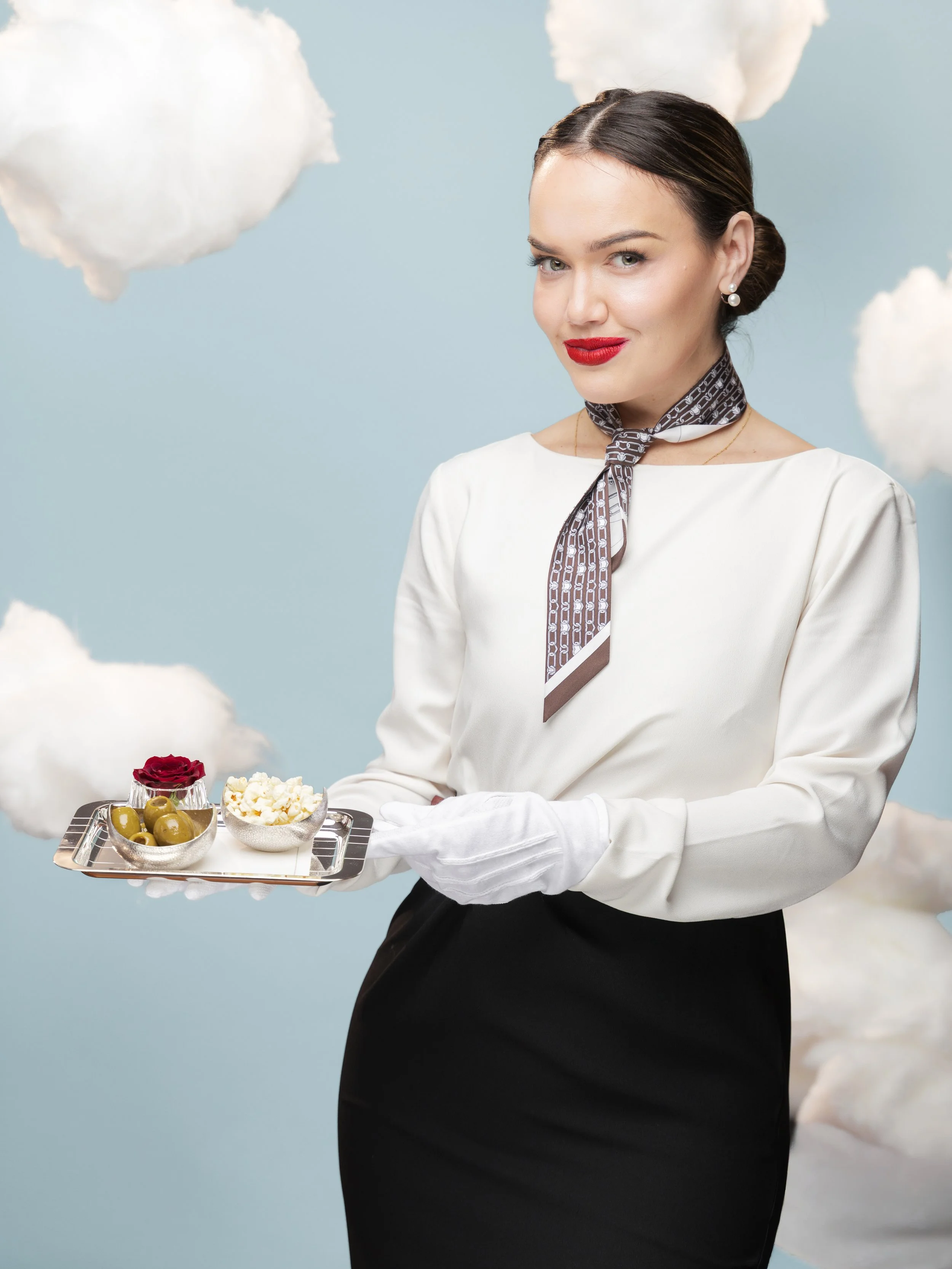 A woman dressed as a flight attendant holding a tray with olives, popcorn, and a red rose, standing against a light blue background with white clouds.