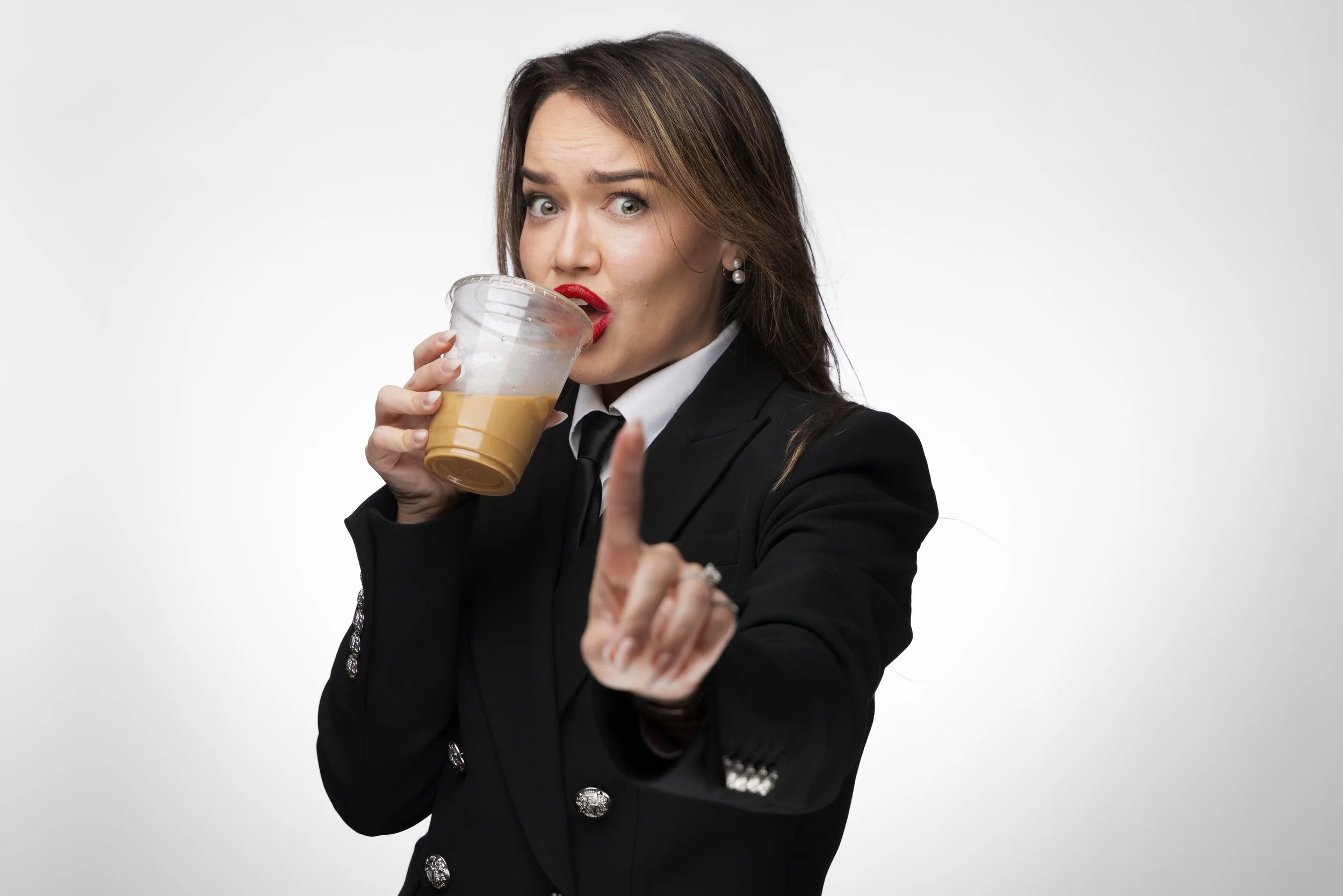 Woman in a black blazer with silver buttons, holding a cup of coffee and gesturing with her finger, with a surprised or serious expression on her face.