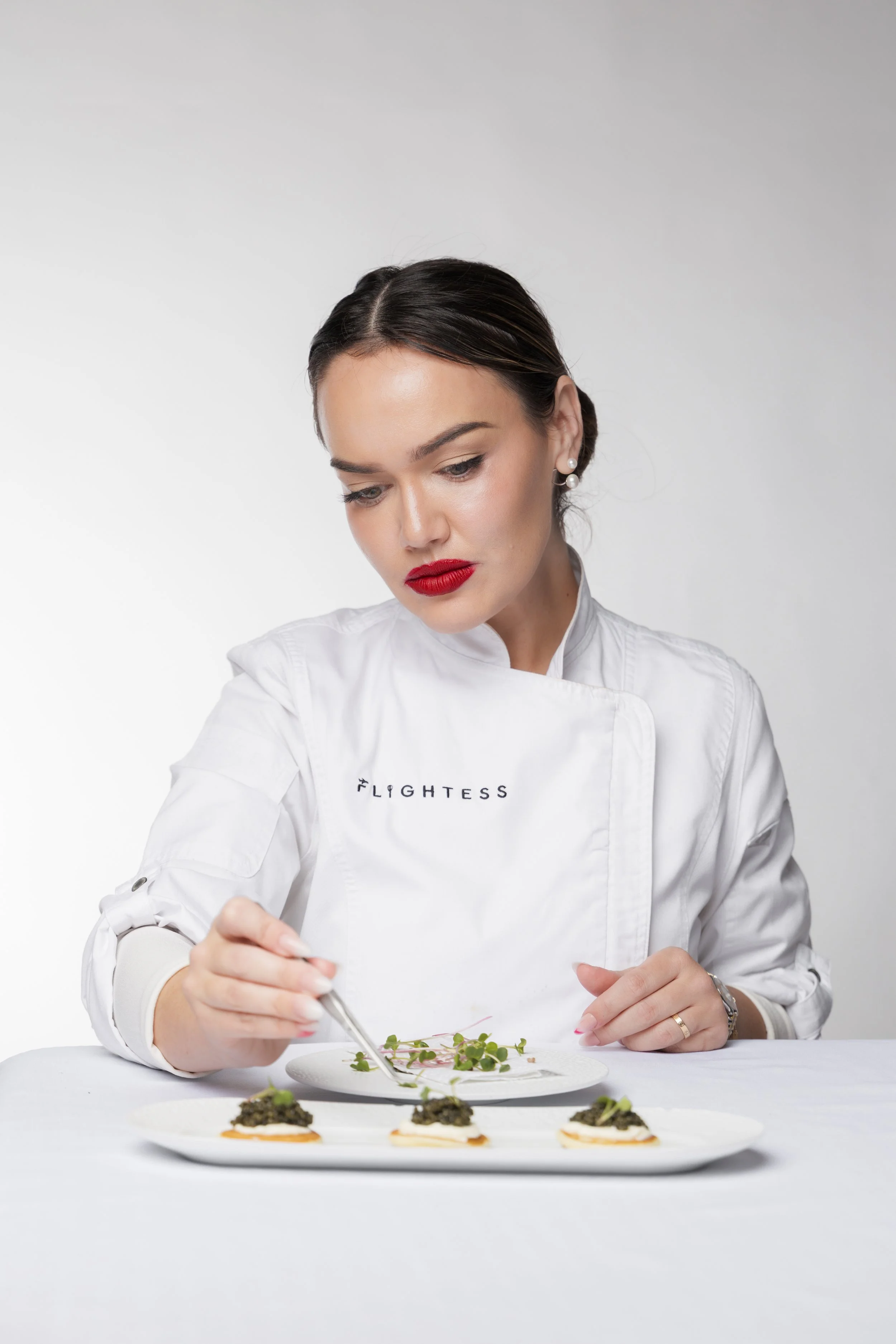 A female chef with dark hair, red lipstick, and pearl earrings, wearing a white chef's coat that says 'Lightess,' garnishes a plate of food with small greens on a white table against a white background.