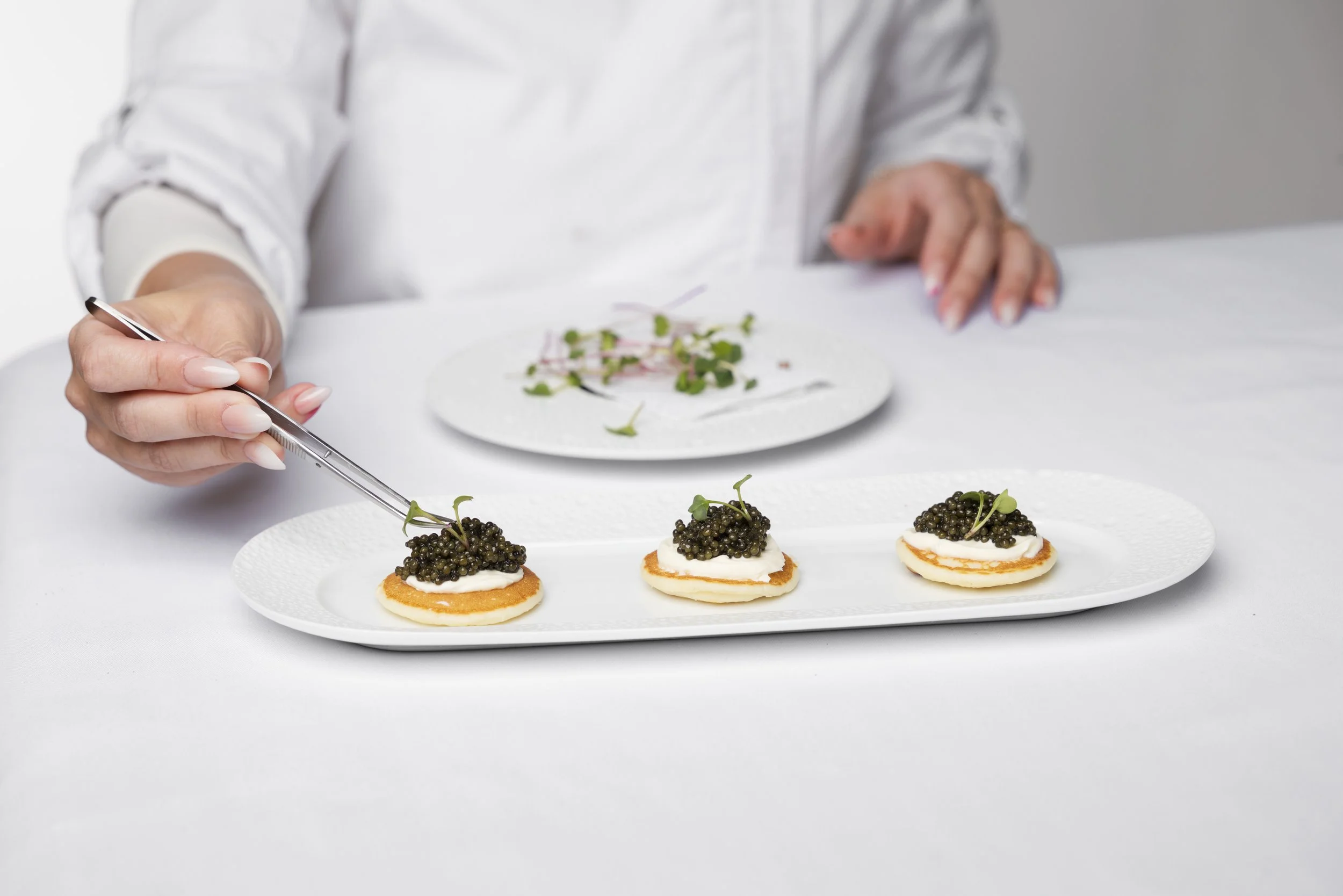 A chef garnishing canapés with caviar and microgreens on a white plate.