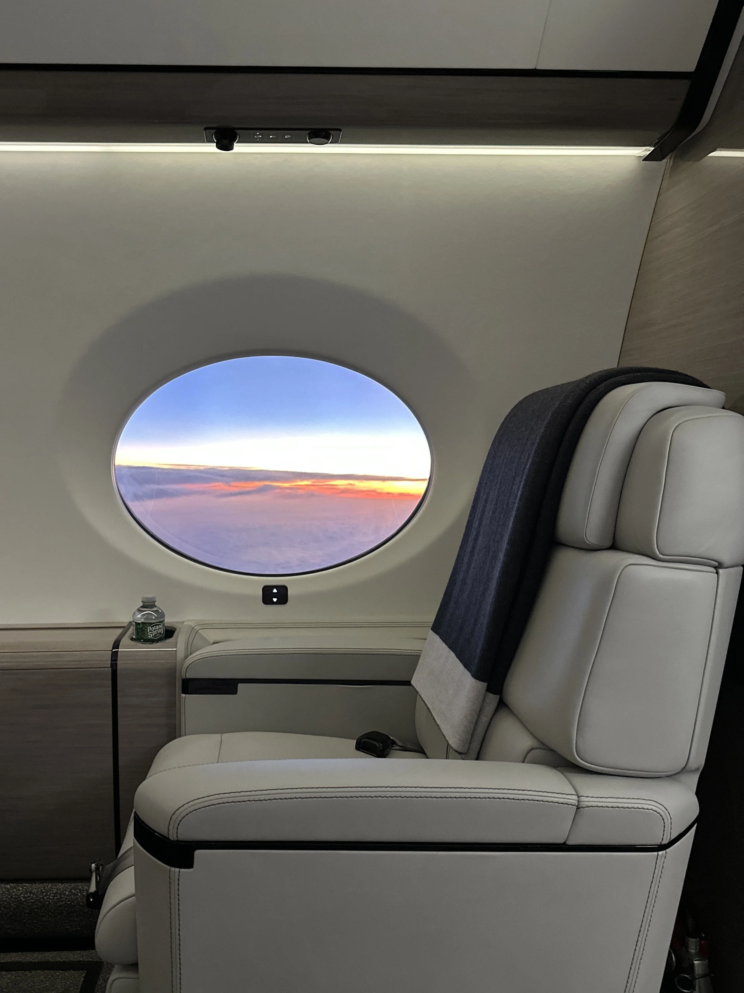 Interior of an airplane cabin with a single business class seat, window view of the sky and clouds during sunset or sunrise, water bottle on a side table, and a blanket on the seat.
