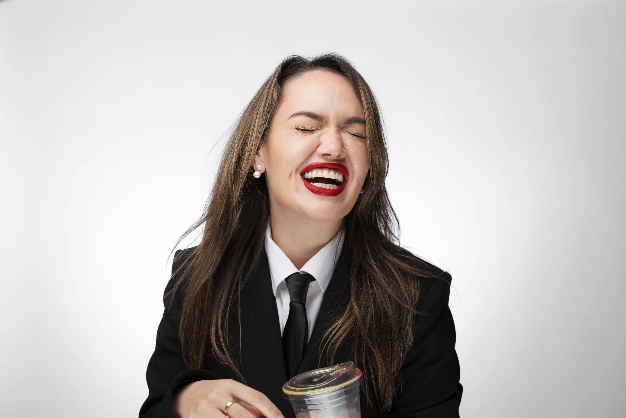 Woman with dark hair wearing a black suit, white shirt, and black tie, laughing with her eyes closed while holding a clear plastic container with a lid.