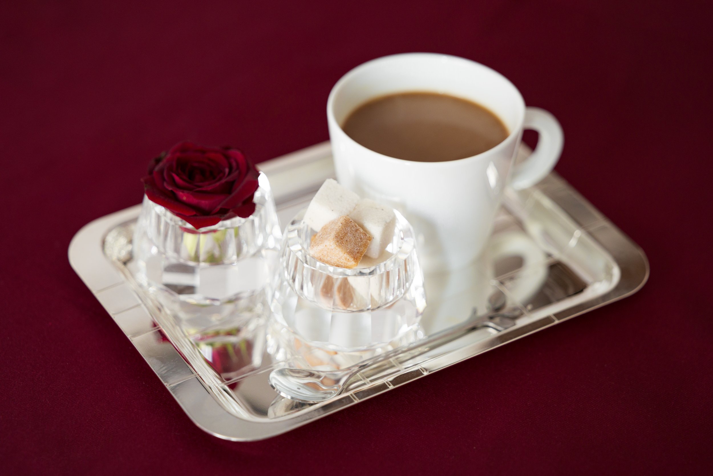 A silver tray with a white cup of coffee, a rose in a glass, and sugar cubes on a dark red surface.