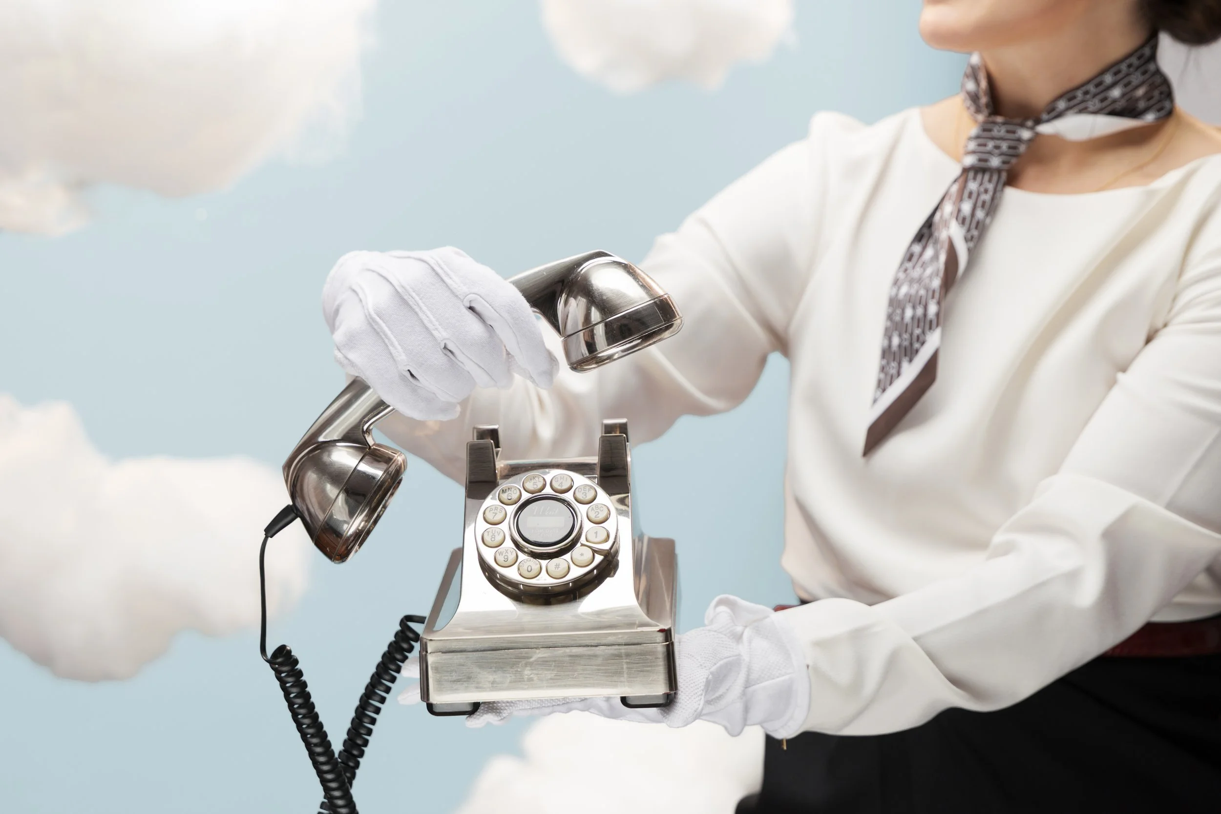 Person wearing white gloves and a white blouse, holding a vintage rotary telephone with a metal base, against a sky with clouds.