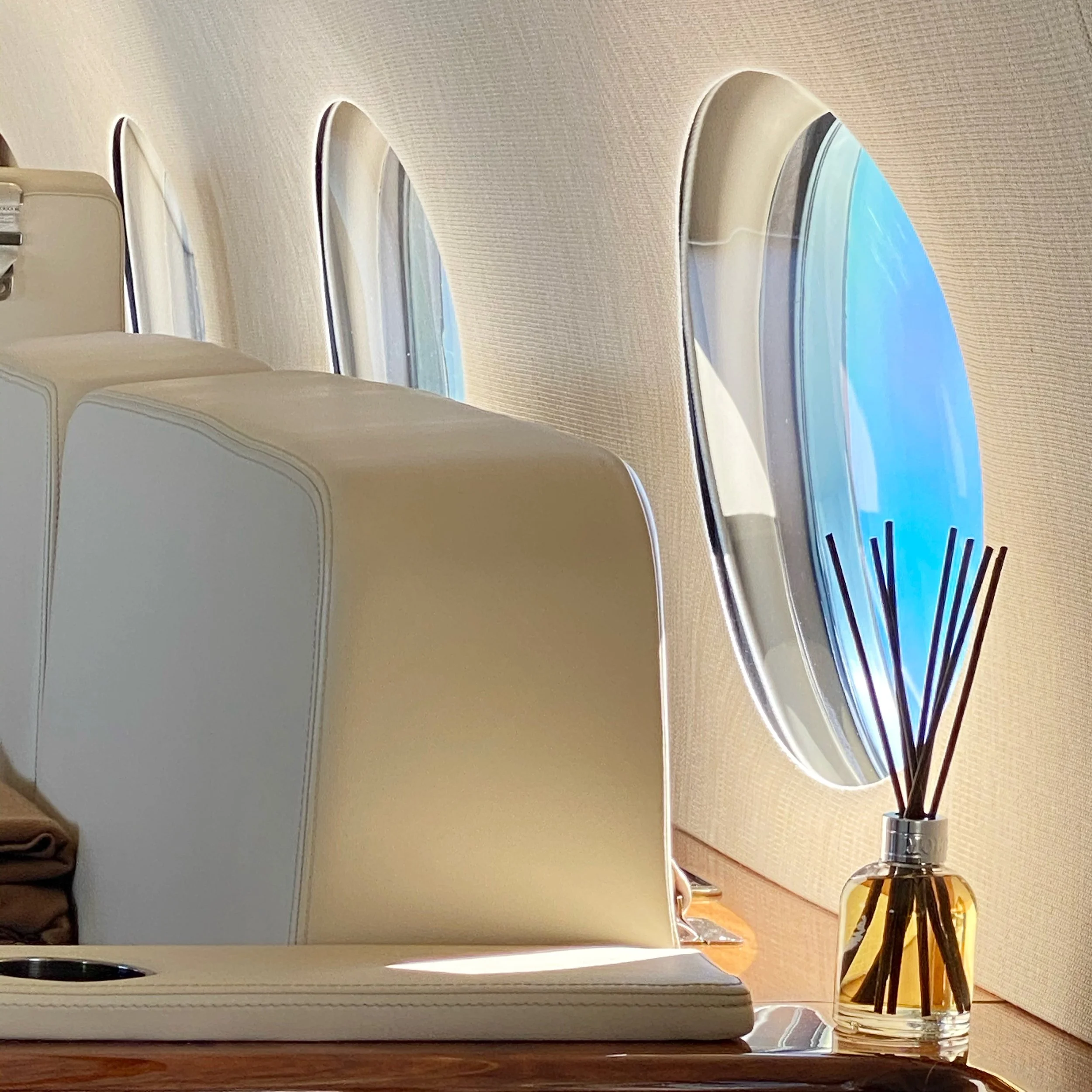 The interior of an airplane cabin, showing several oval airplane windows with a view of blue sky and clouds. There is a small table with a decorative reed diffuser in a glass bottle.