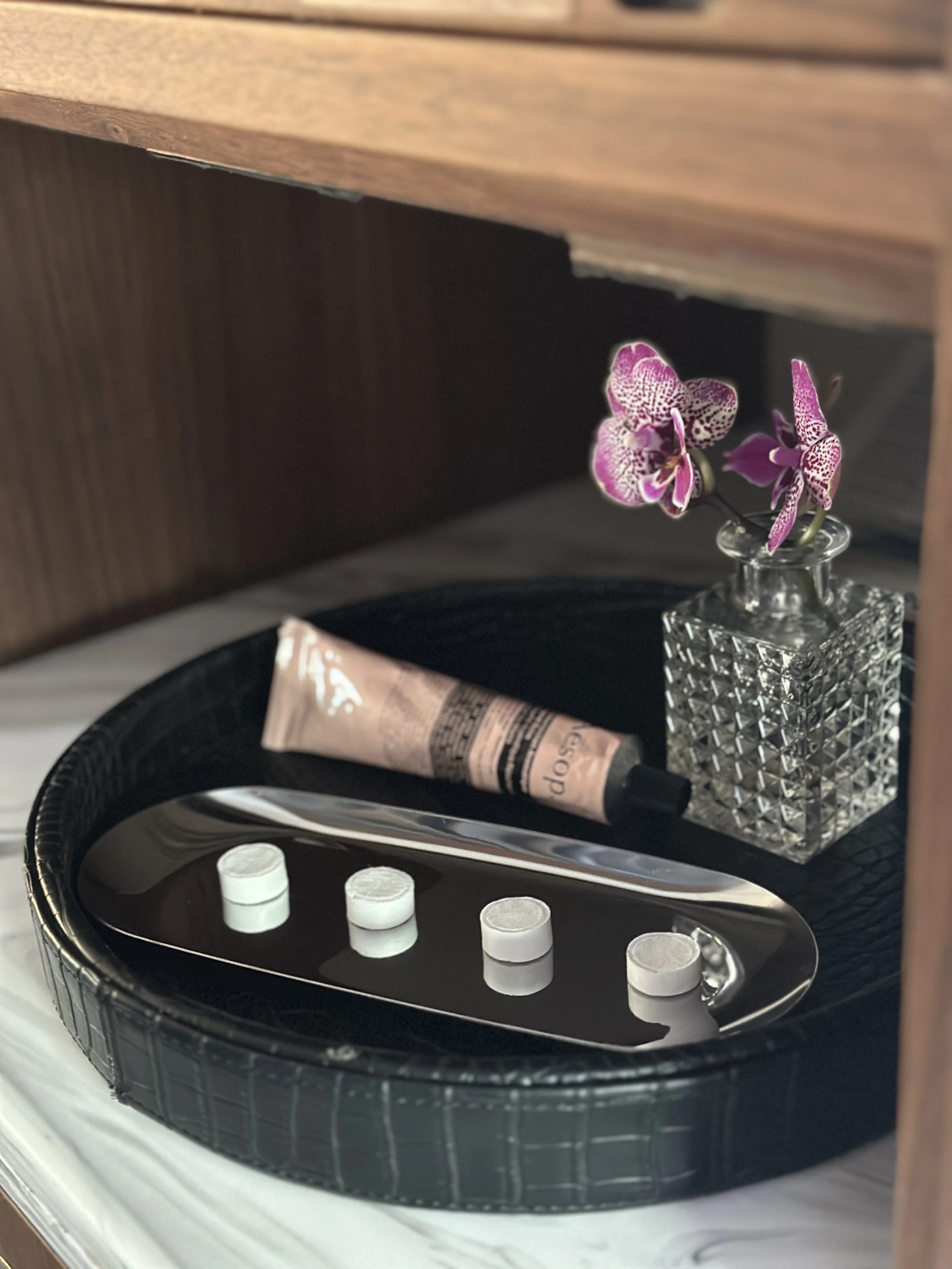 A black tray with four white tablets, a silver spatula, a pink tube, and a glass vase with pink and purple orchids.
