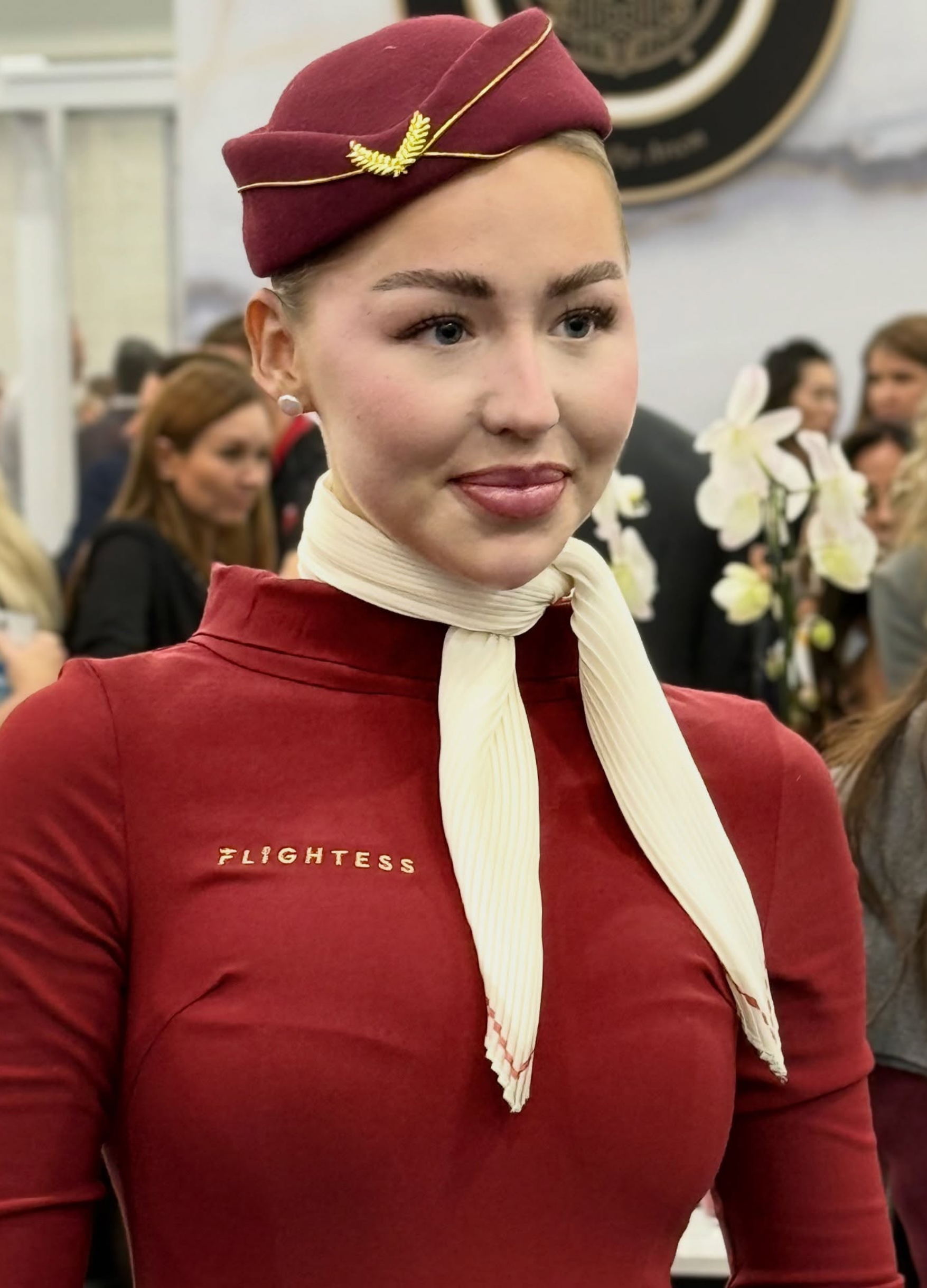 A woman dressed as a flight attendant against a background of people and white orchids, wearing a maroon hat with a gold decoration, a maroon uniform with the word "FLIGHTESS" on it, and a cream-colored neck scarf.