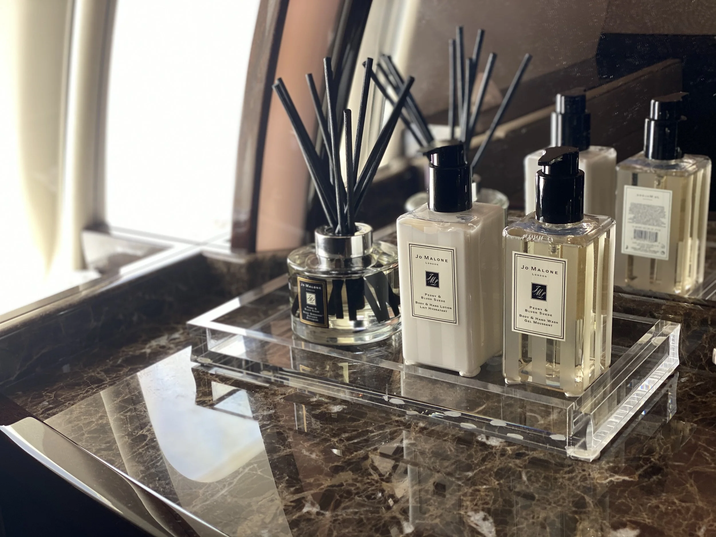 A clear acrylic tray on a dark marble countertop holds three bottles of Jo Malone body lotion and body wash, along with a diffuser with black reeds. A mirror and a window are reflected in the background.