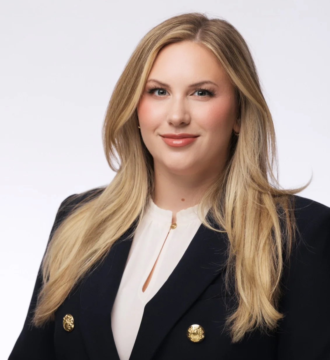 Professional woman with long blonde hair, fair skin, wearing a black blazer with gold buttons and a white blouse, smiling at the camera, against a plain light gray background.