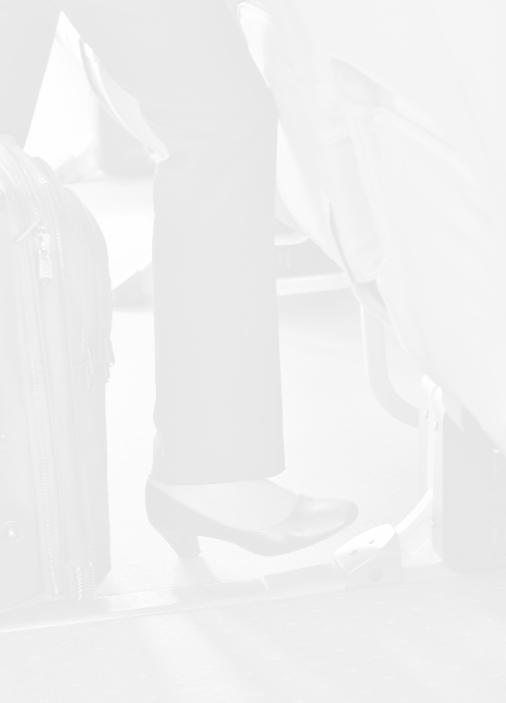 Close-up of a passenger's leg and foot as they sit on an airplane, wearing black dress shoes and dark pants, with a rolling suitcase beside them.