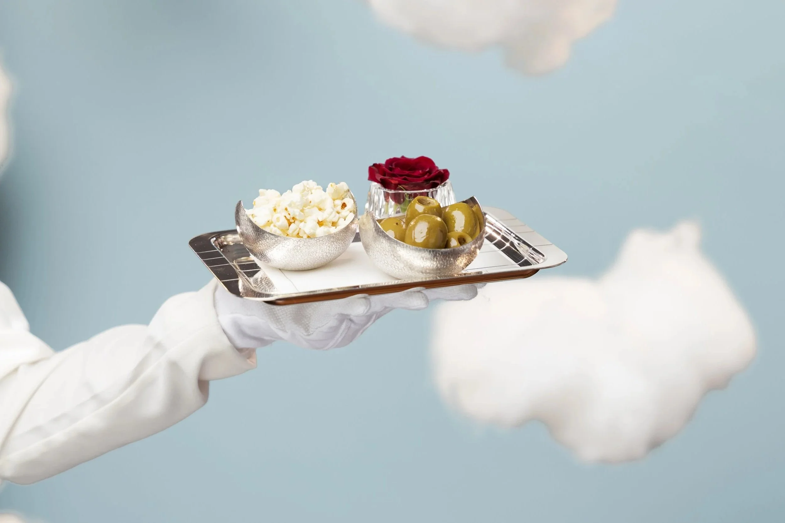 Person wearing white gloves holding a tray with silver bowls of popcorn, green olives, and a glass of red rose flower, against a cloudy sky background.