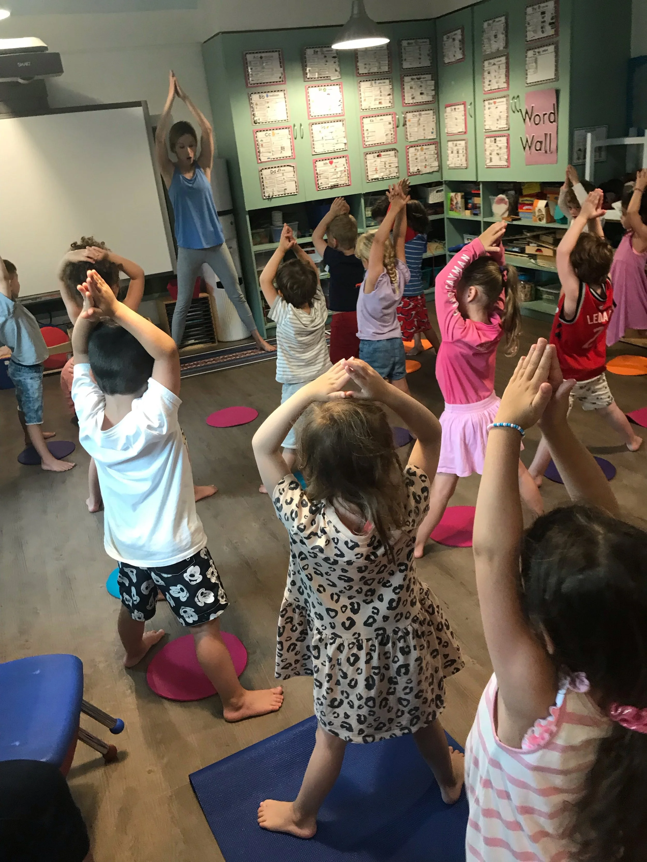 Children in a classroom participating in an exercise activity led by a young instructor standing on a platform.