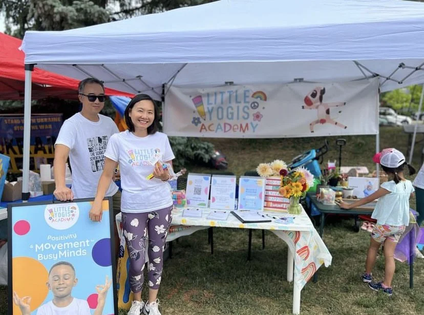 Two women and a girl at a Little Yogi Academy outdoor booth with colorful sign and table, under white canopy tent, on grass, with trees and other tents in background.