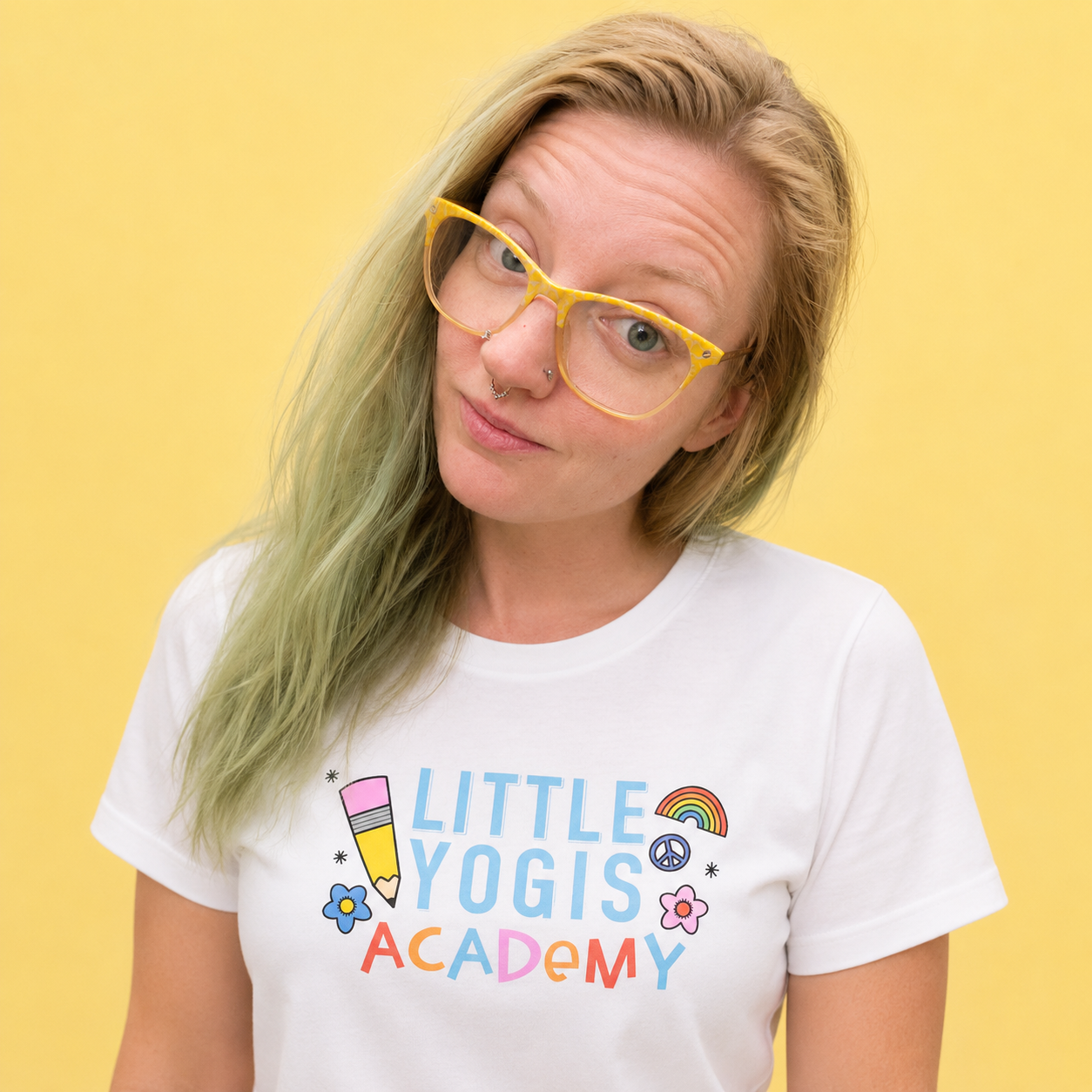 A woman with long blonde hair, wearing yellow glasses and a white T-shirt with colorful illustrations and text reading 'Little Yogis Academy,' standing against a yellow background.
