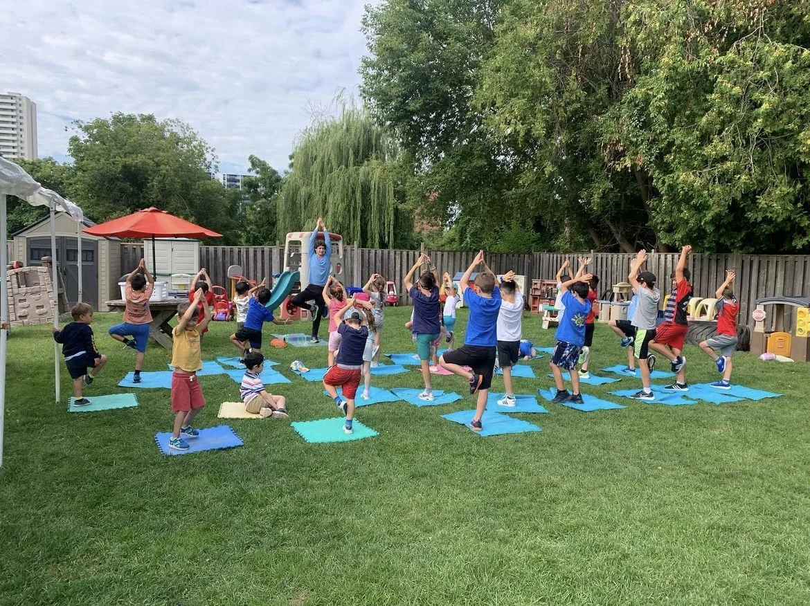 Children doing outdoor yoga on mats in a backyard with trees, a slide, and a wooden fence