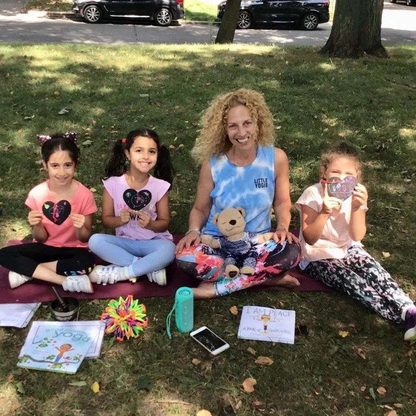 Group of four women and children sitting on a blanket in a park, holding handcrafted heart-shaped cards with peace symbols. There are colorful items, a microphone, a phone, and a sign that says "I AM PEACE" on the ground.
