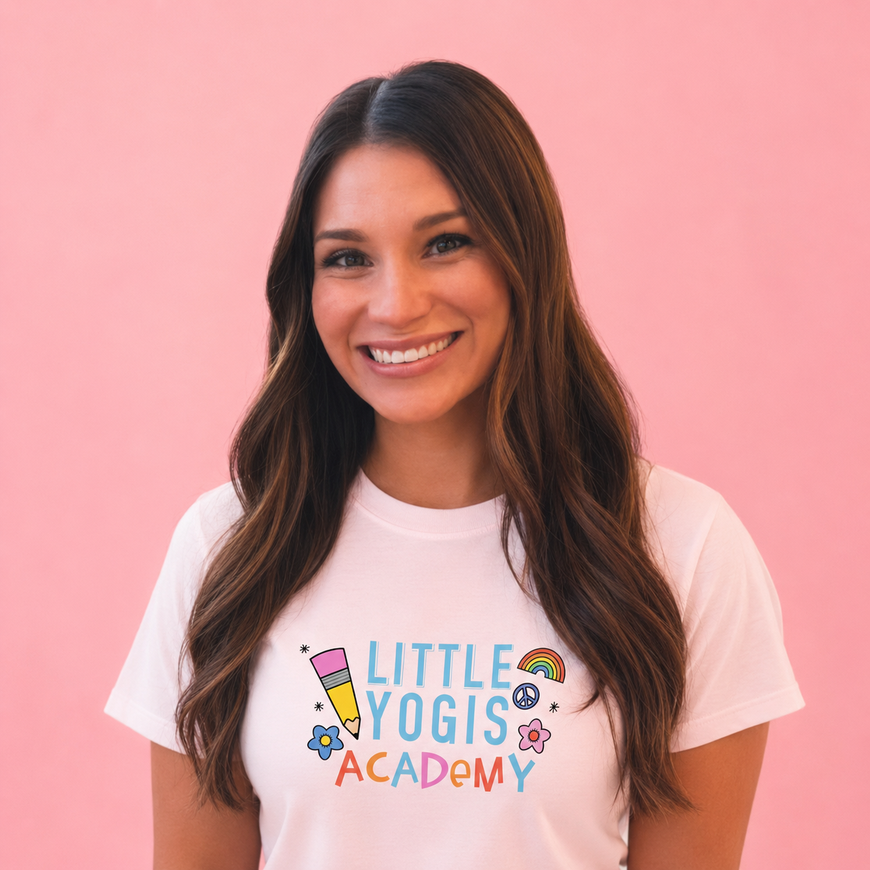 A smiling woman with long brown hair against a pink background, wearing a white T-shirt with colorful text and illustrations that reads 'Little Yogis Academy'.