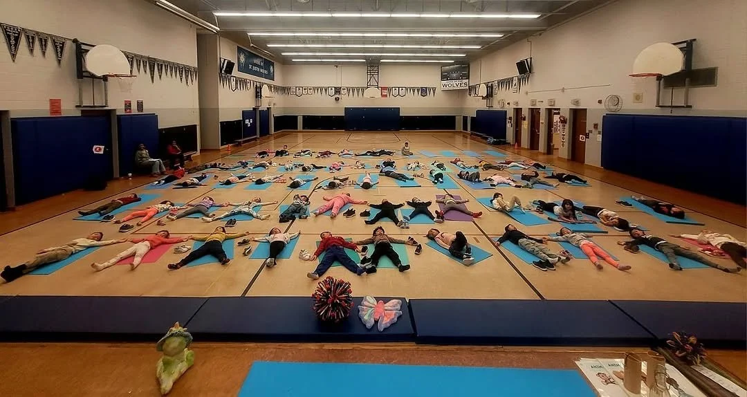 Children participating in a group exercise class in a gymnasium, lying on blue mats in a starfish position.