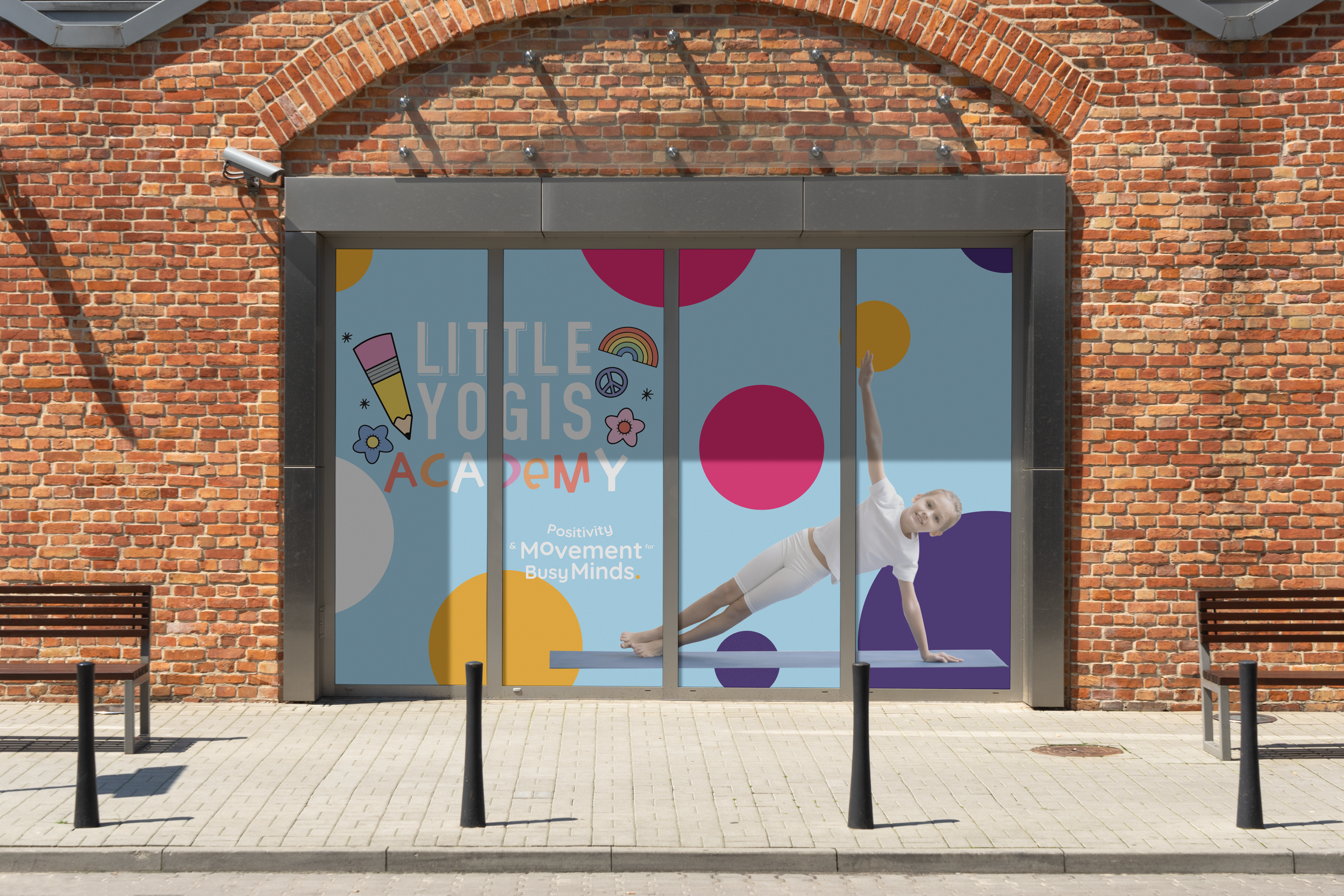 Storefront window with a large illustration for Little Yogis Academy, featuring a young girl doing a yoga pose on a purple yoga mat, surrounded by colorful circles and playful graphics, with the storefront made of red brick.