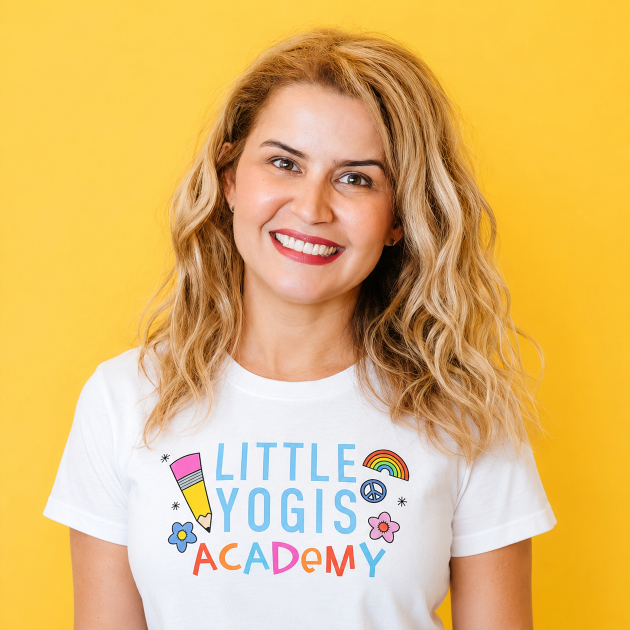 A woman with blonde curly hair smiling, wearing a white t-shirt that says Little Yogis Academy with colorful graphics, standing against a yellow background.