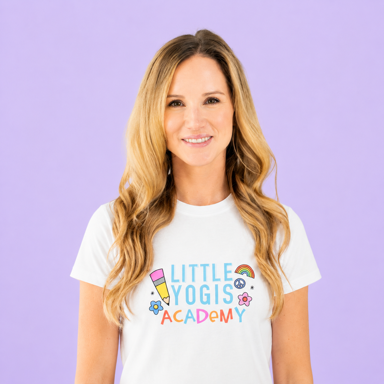 A woman with long wavy blonde hair smiling at the camera wearing a white T-shirt with colorful graphics and text that reads 'Little Yogis Academy' against a purple background.