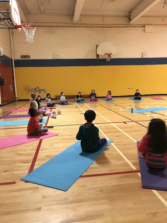 Children practicing meditation or yoga on colorful mats in a gymnasium.