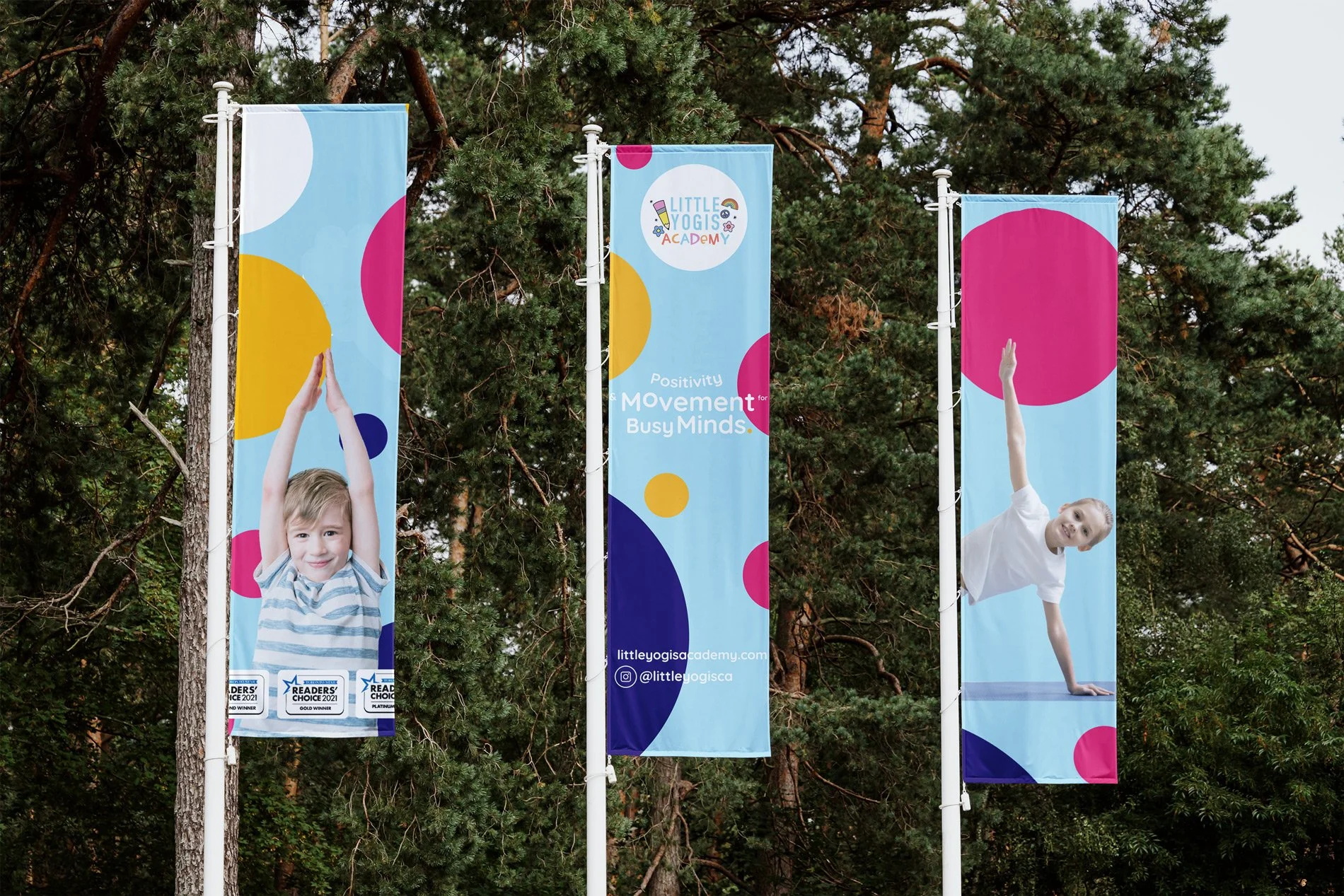 Three colorful vertical banners hanging among trees. The middle banner displays the text 'Little Yogis Academy' and 'Positivity Movement Busy Minds,' along with social media info. The left and right banners feature children performing yoga poses, smi