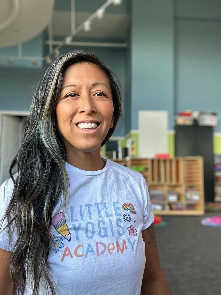 A smiling woman with long, dark hair and gray highlights, wearing a white t-shirt with colorful text and illustrations, standing indoors in a colorful room.