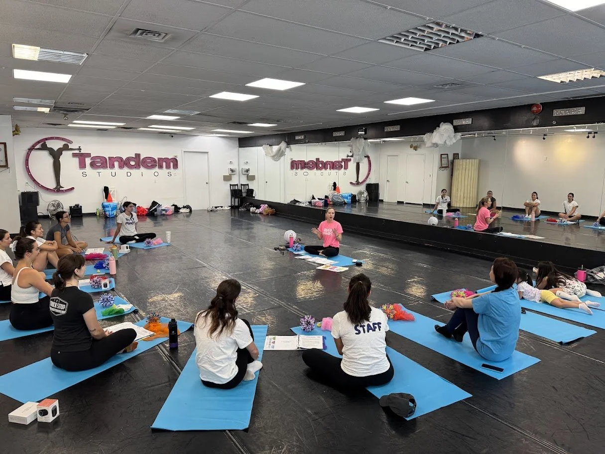 A group exercise class at Tandem Studios, with participants sitting on blue mats on the floor while an instructor leads a session in front of a mirror. The studio has a white wall with the Tandem Studios logo and a large mirror along one side.