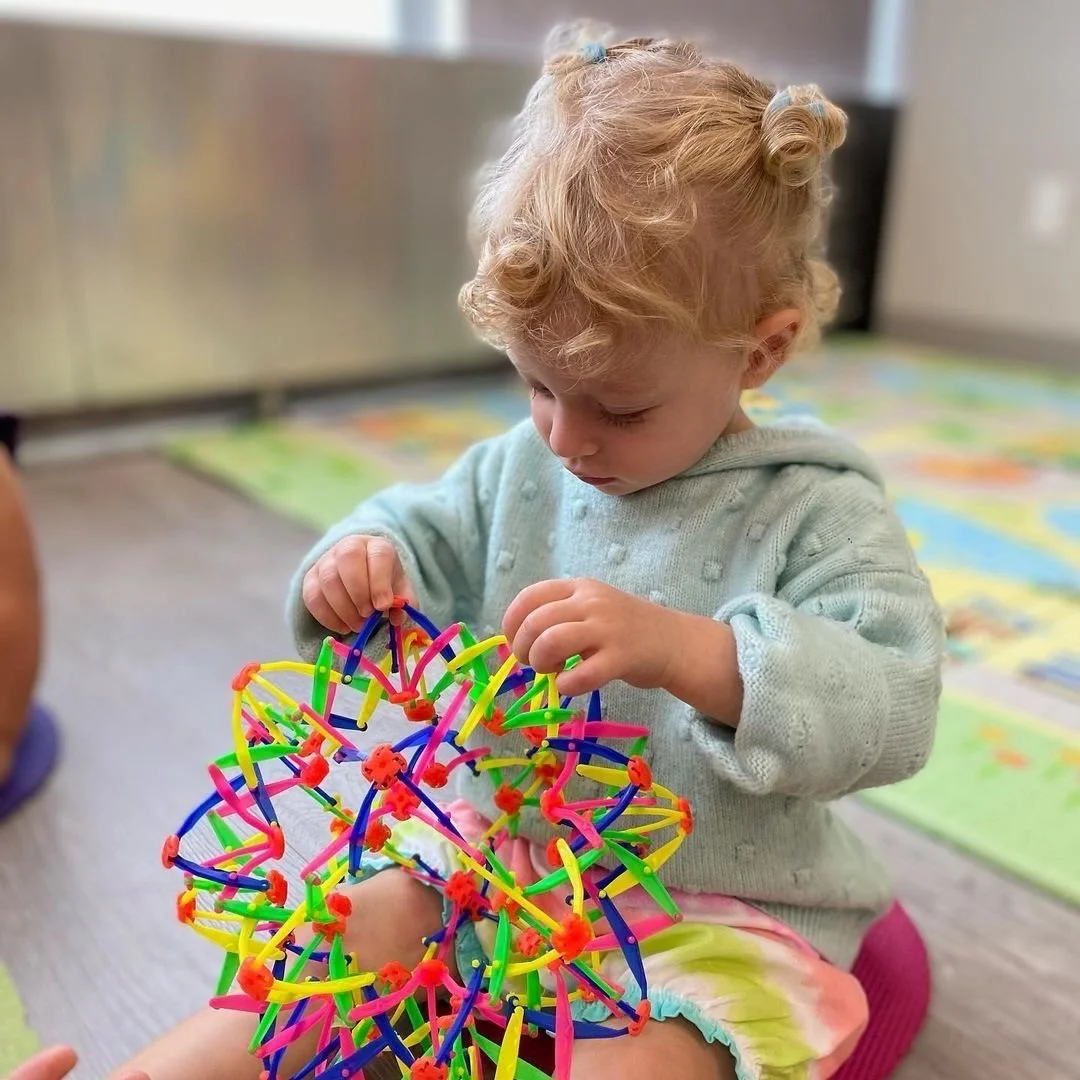 A young girl with curly blonde hair playing with a colorful, plastic thimble toy, sitting on a colorful play mat.