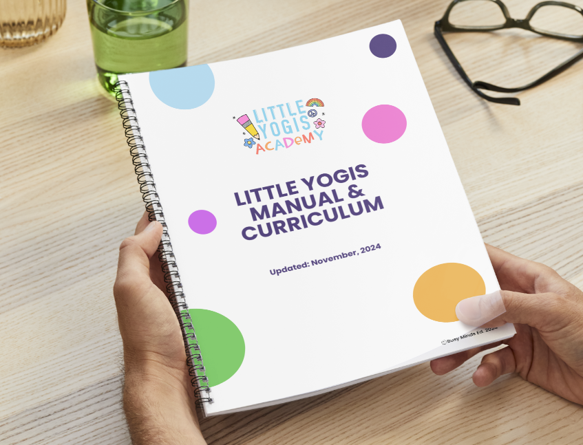 A person holding a spiral-bound manual titled "Little Yogi's Manual & Curriculum" with colorful dots on the cover, sitting on a light wooden table with a glass of water, glasses, and a pen nearby.