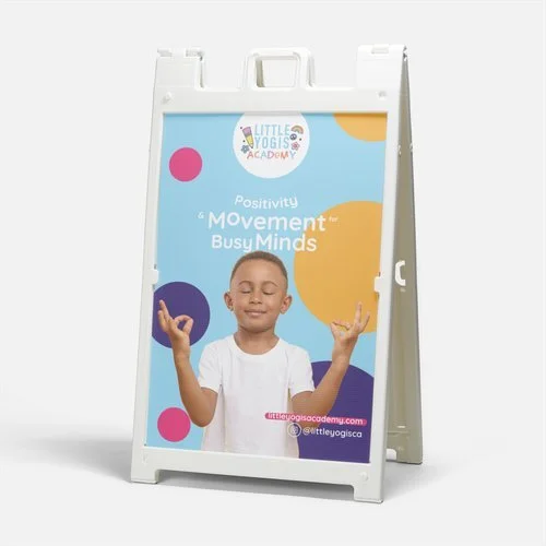 A white A-frame sign board with a colorful poster from Little Yogis Academy promoting positivity and movement for busy minds, featuring a smiling child with closed eyes and hands in a calming gesture.