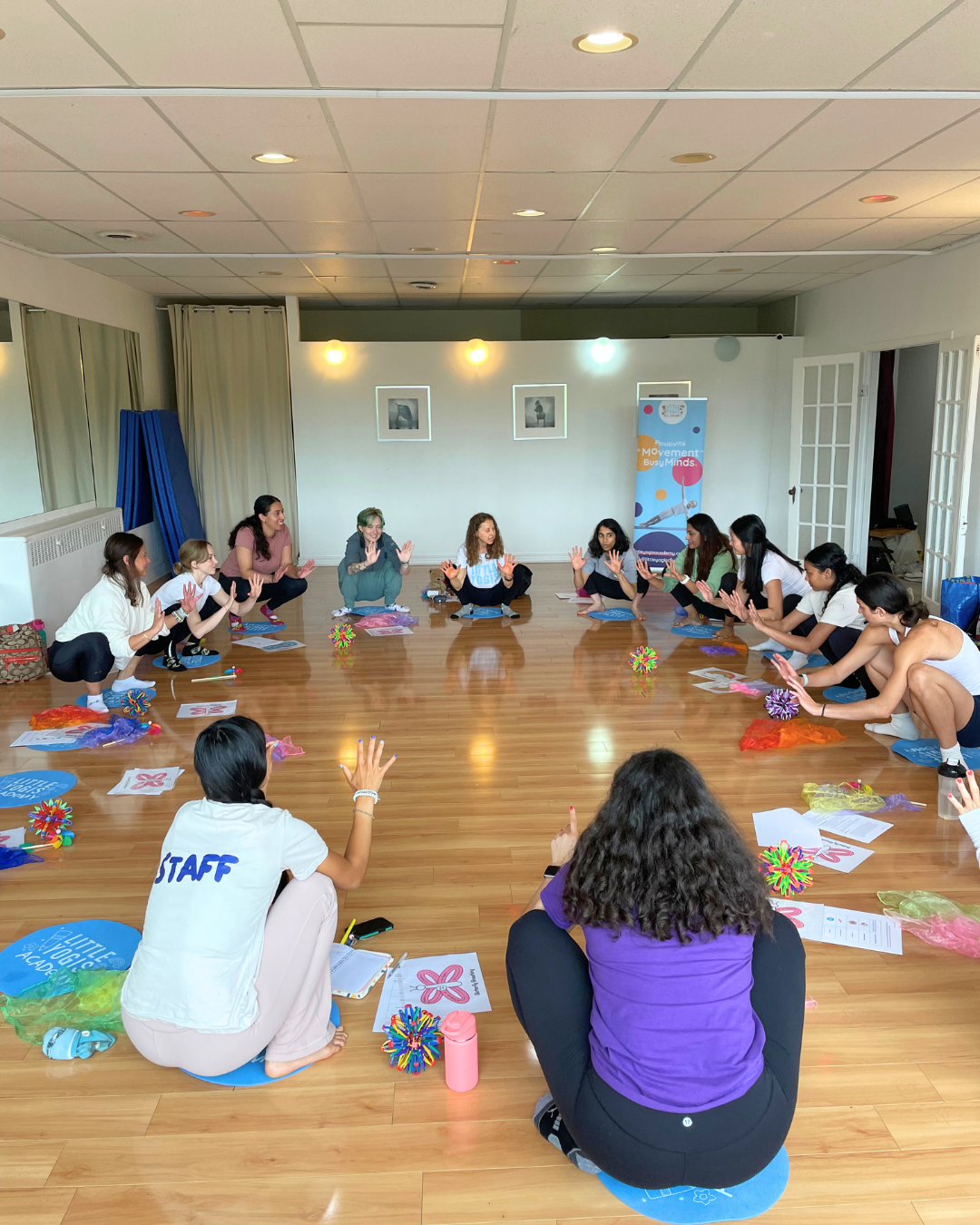 A group of women and girls participate in a seated circle activity indoors, with some raising hands, on a wooden floor with colorful props and papers, in a well-lit room with artwork on the wall and a banner reading 'Positive Movement Bus Minds'.