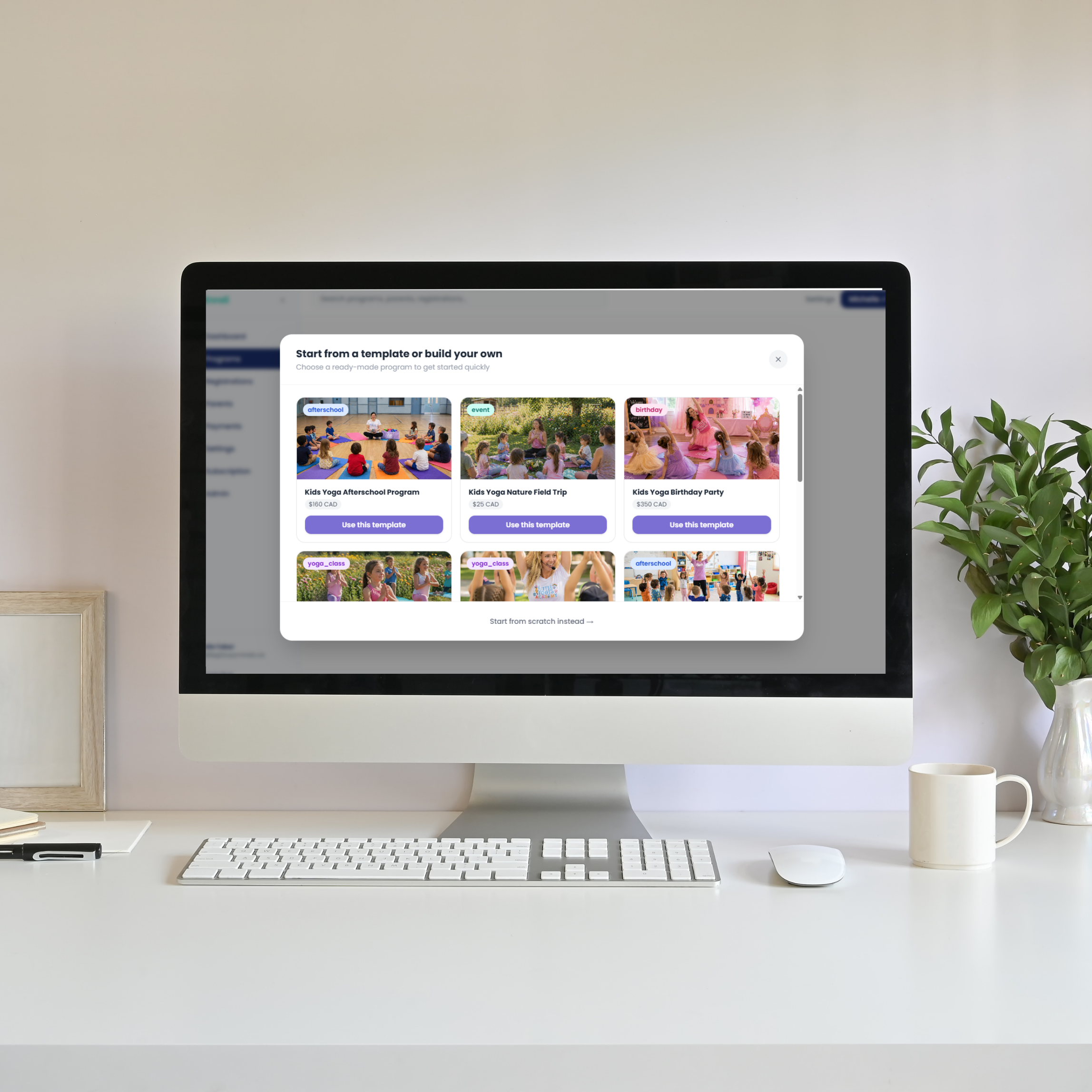 A clean white desk with an iMac computer displaying a webpage about children's activities, a white keyboard, a mouse, a white cup, a black pen, a small white stack of paper, a potted plant, and a wooden picture frame on the left side.