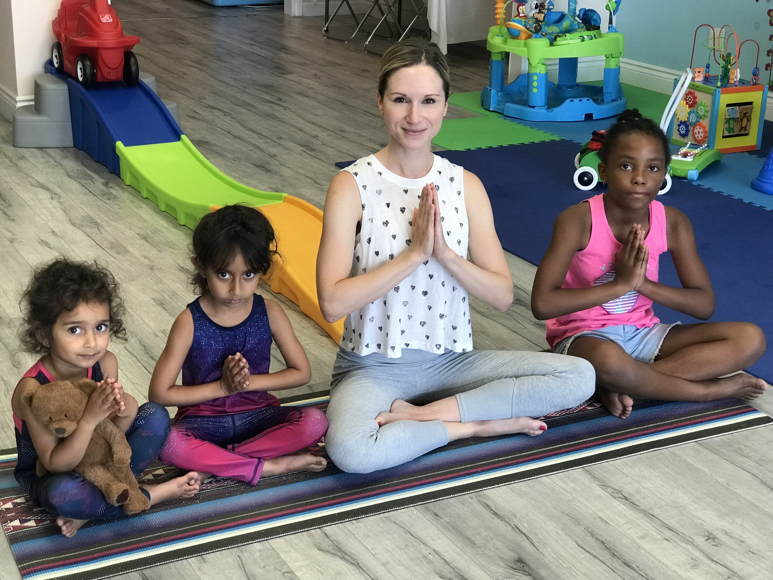 Four children and a woman in a yoga pose with hands together in a prayer position, sitting on a striped mat in a children's playroom. The children are holding stuffed animals and have serious expressions. The room has colorful play equipment in the background.