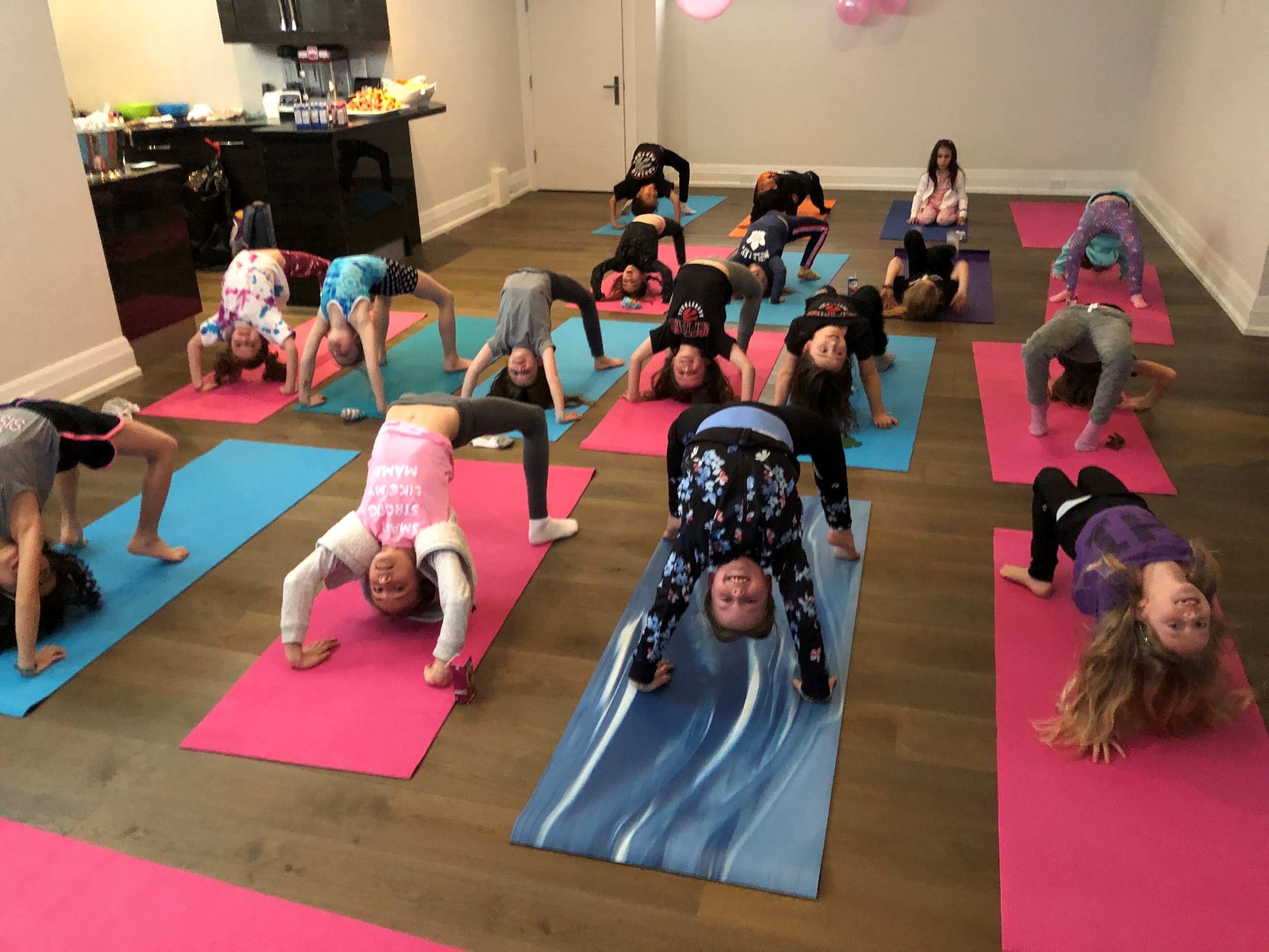 Group of children practicing yoga on colorful mats in a room with hardwood floors.