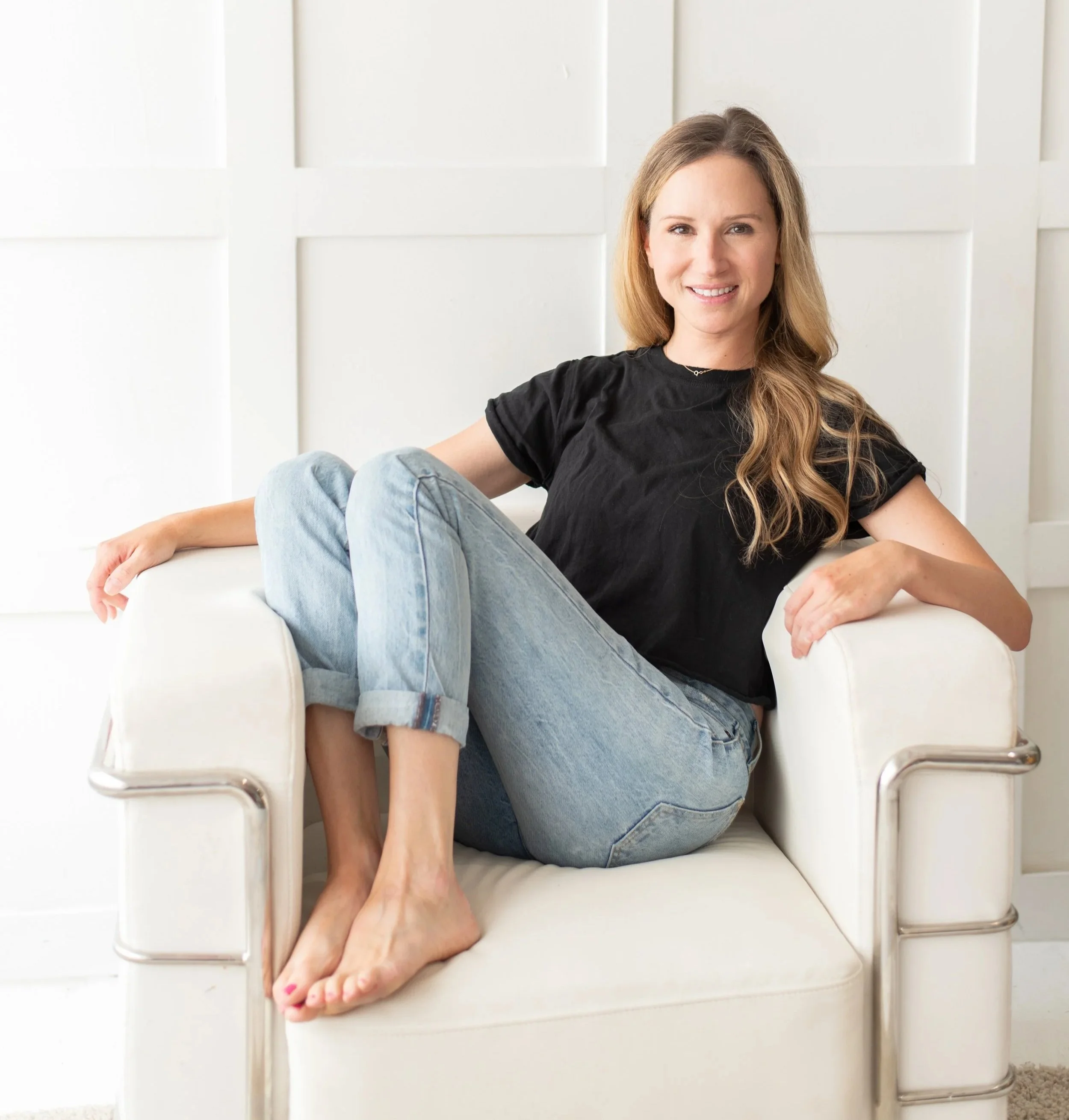 A woman with long wavy hair sitting comfortably on a white armchair in a minimalist room with white walls, wearing a black t-shirt and light blue jeans, smiling at the camera.