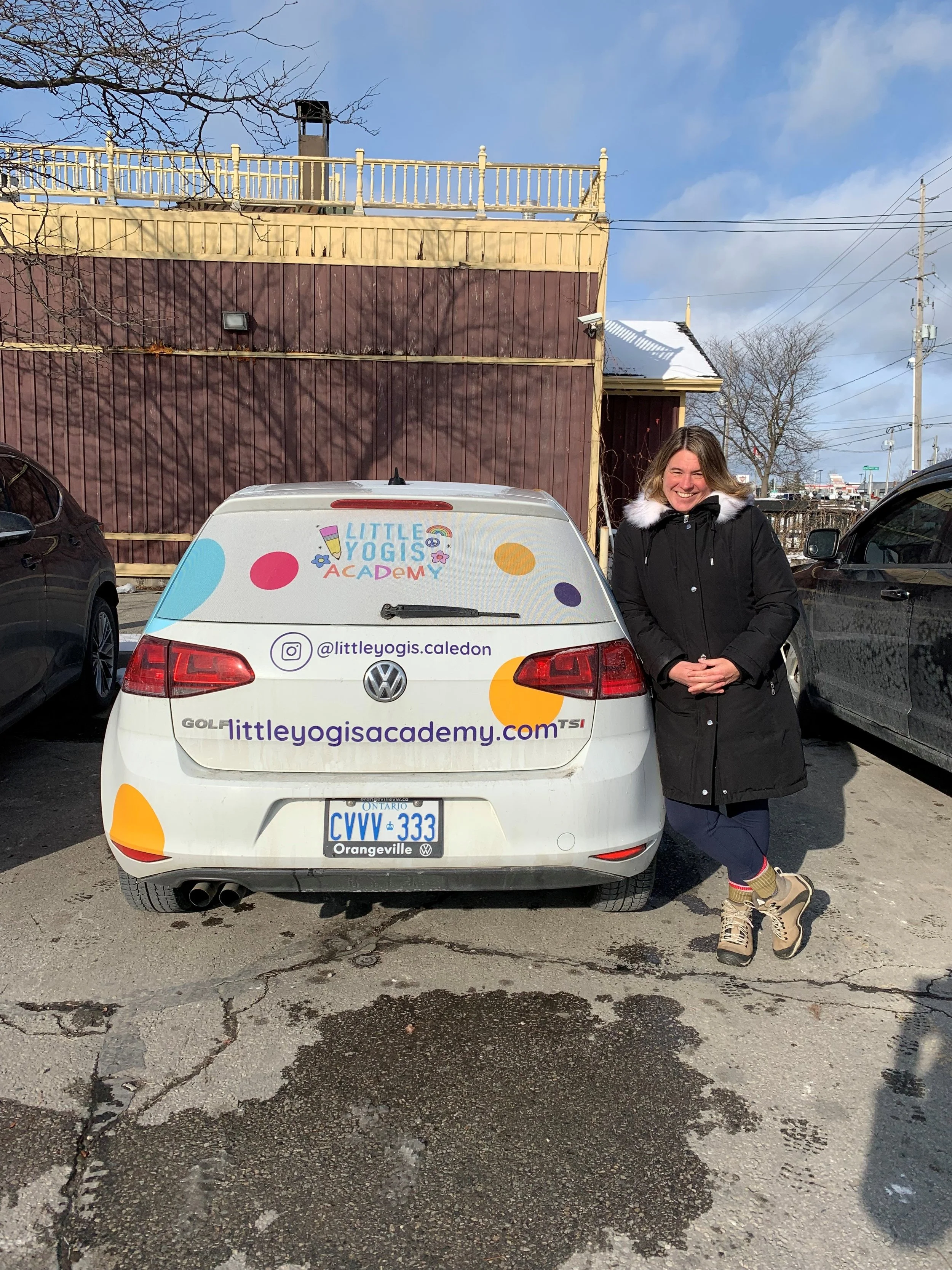 A woman standing next to a white Volkswagen car with Little Yogis Academy branding on the back, in a parking lot with a brown building and a clear blue sky.