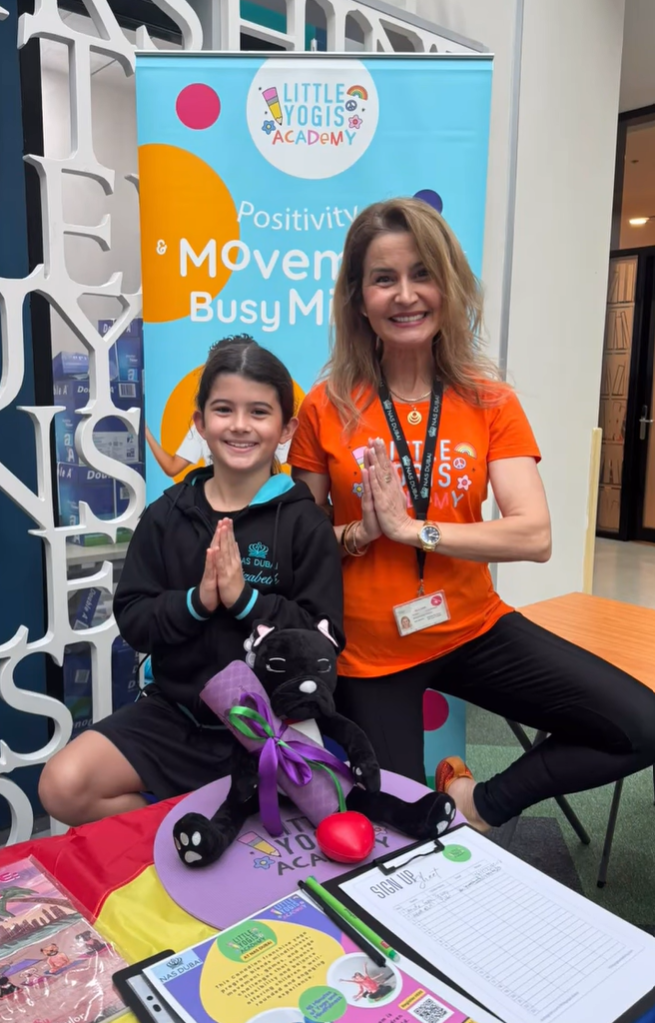 A smiling woman and girl in front of a Meetup banner, both with hands pressed together in a prayer gesture. The girl holds a black stuffed cat with a purple bow and a small red heart in front of her. The table in front of them has colorful papers, a 