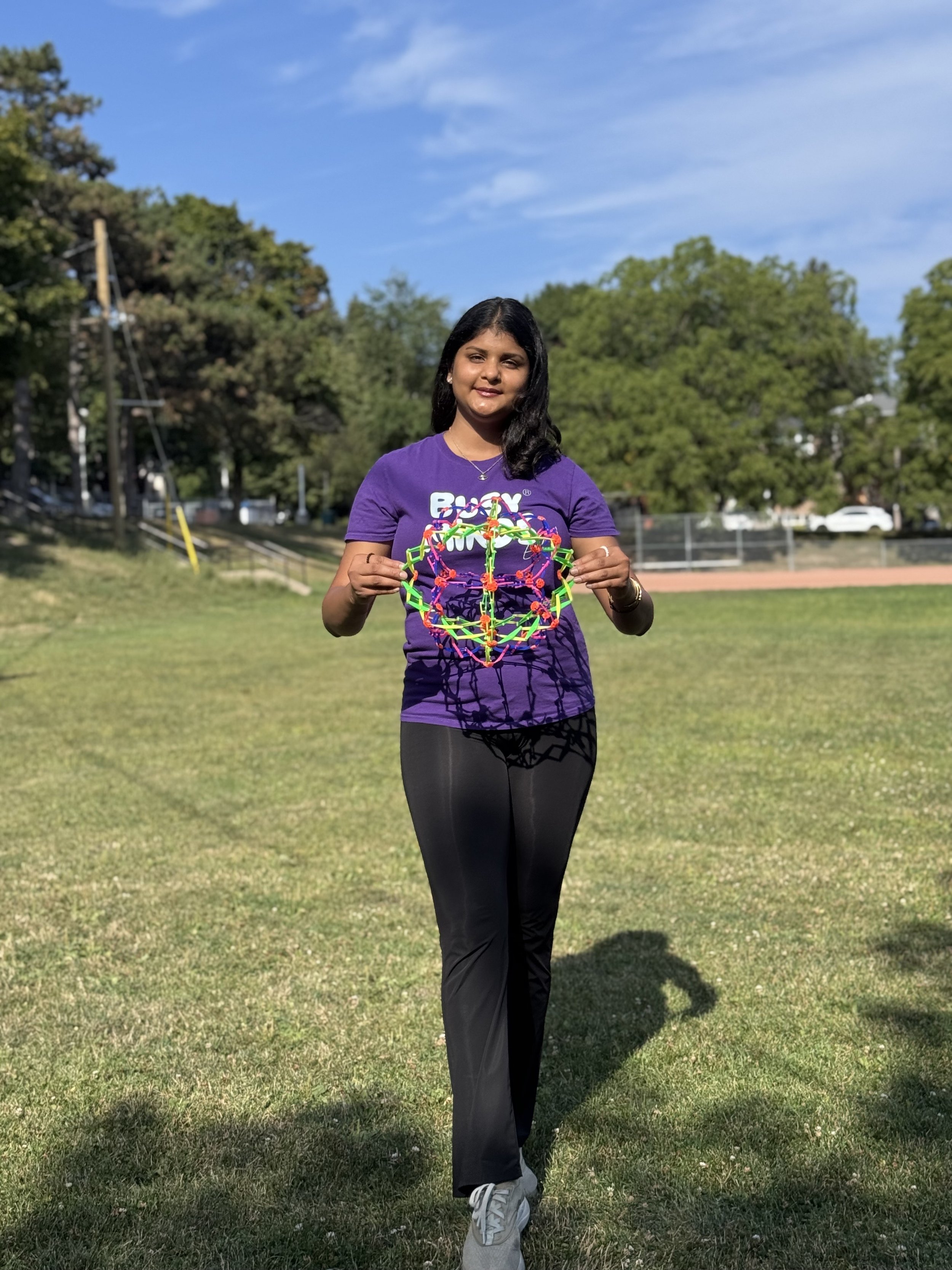 Young woman standing outdoors on grass holding a colorful geometric toy, wearing a purple t-shirt and black pants, with trees and a blue sky in the background.