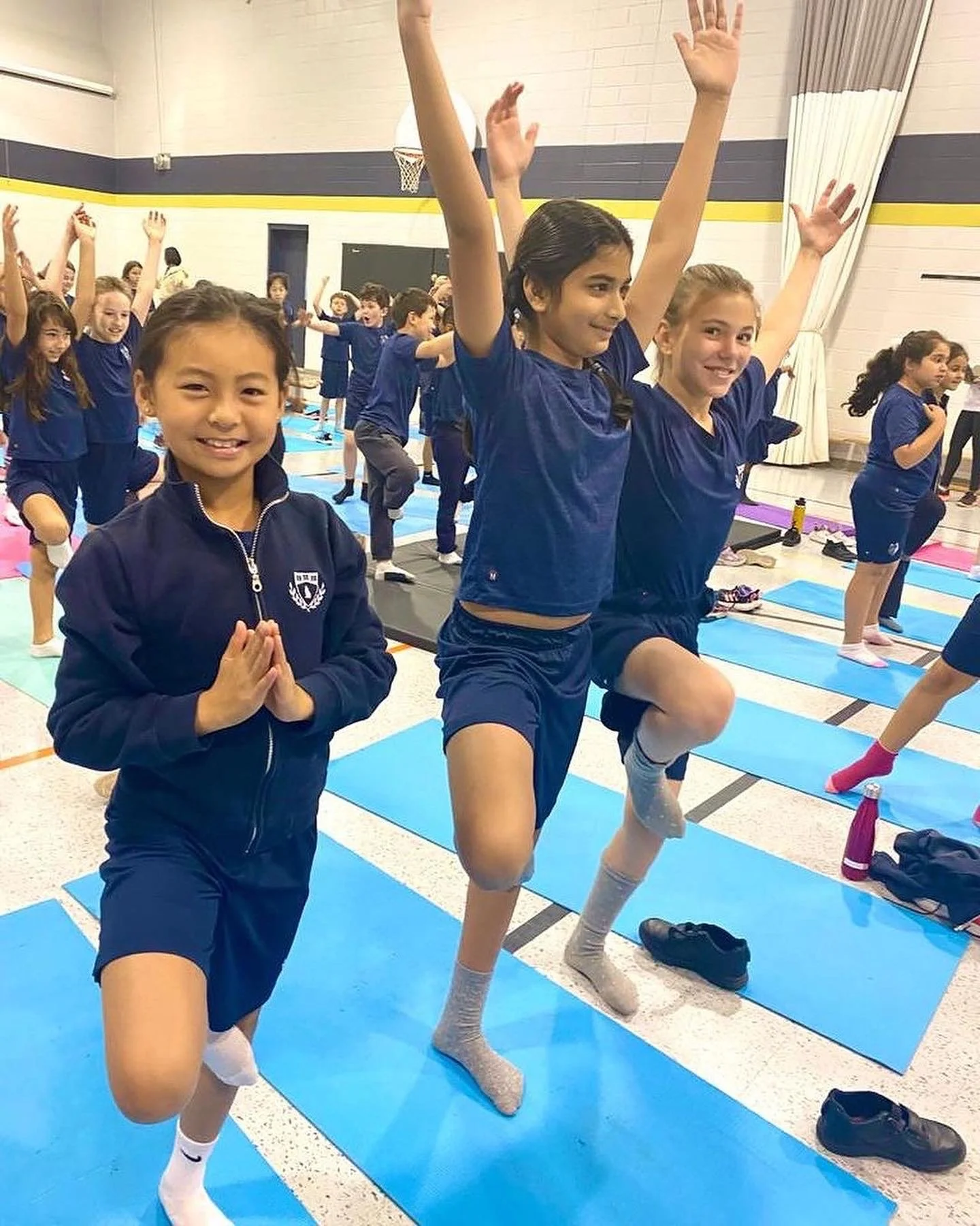 Group of children practicing yoga in a gymnasium, some in tree pose with hands together and others with arms raised, all dressed in blue sports uniforms.