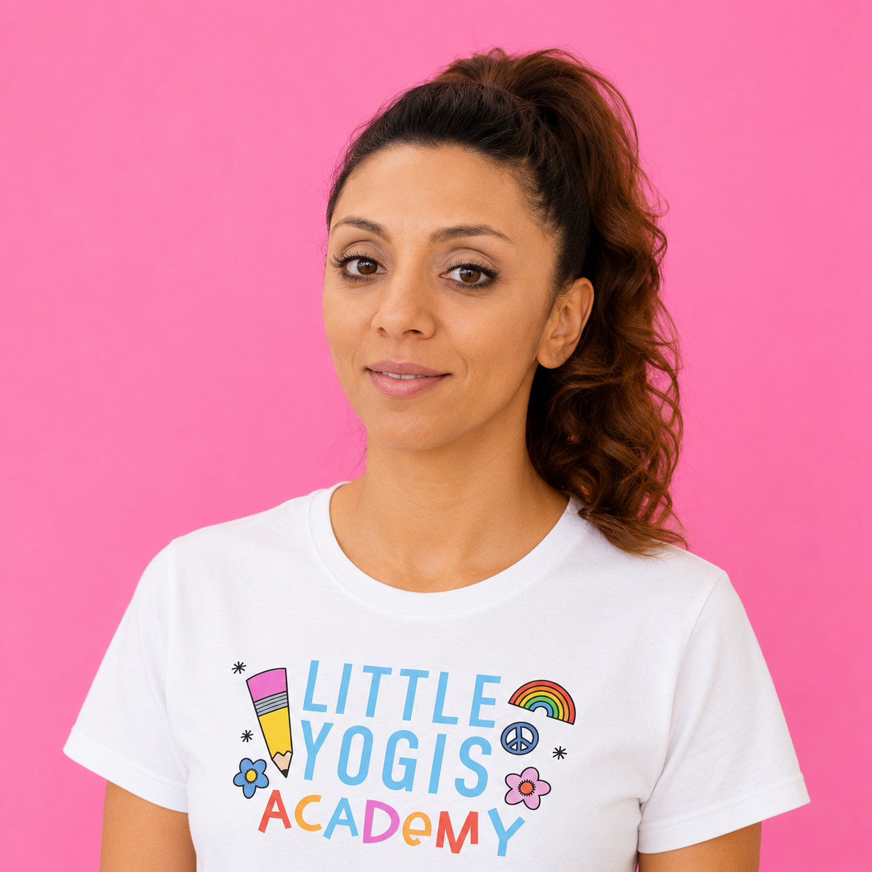 A woman with brown hair in a high ponytail wearing a white T-shirt with colorful text and graphics, standing against a pink background.