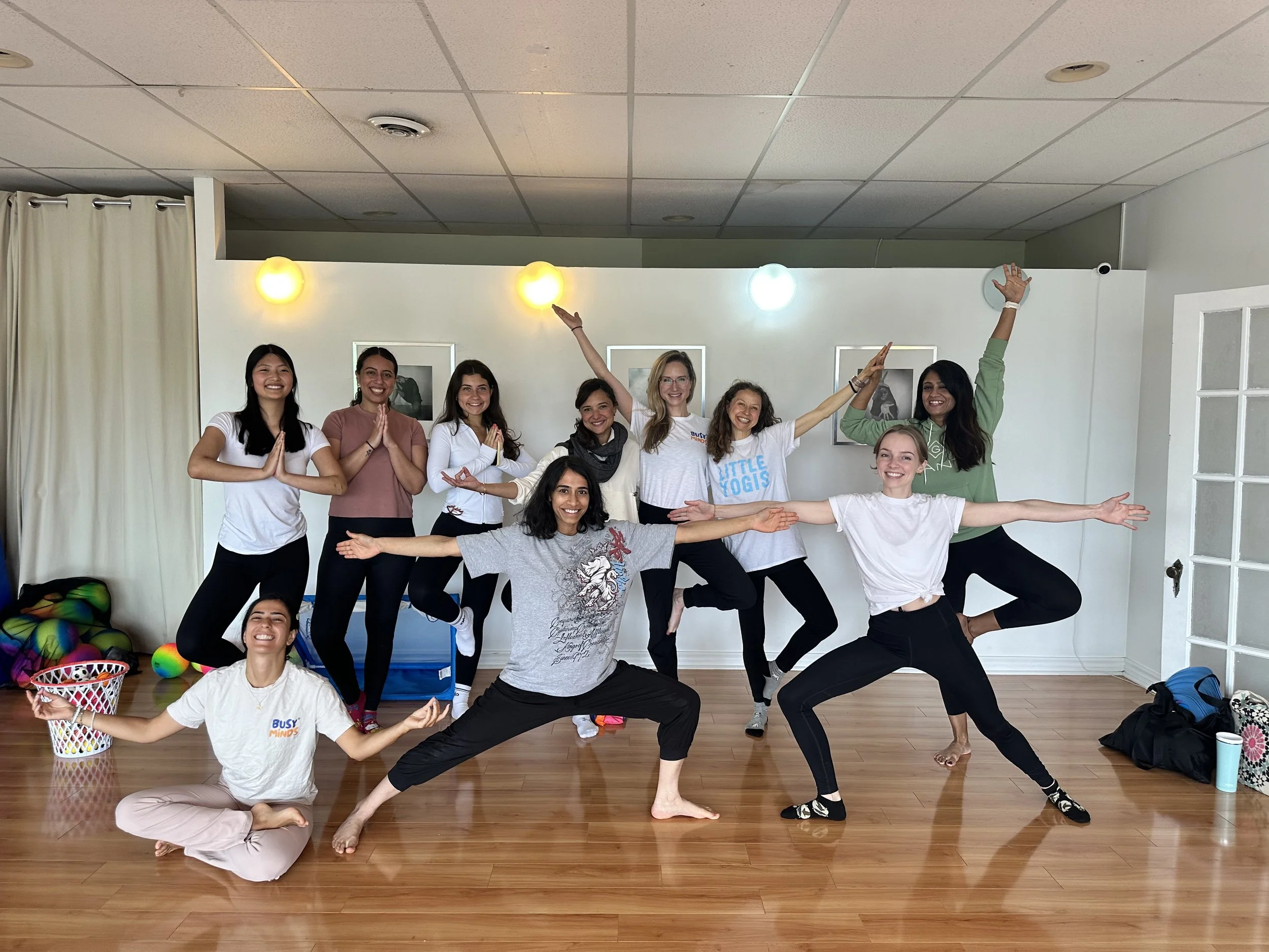 Group of nine women practicing yoga in a spacious indoor room with wooden floors, white walls, and framed art. Some are standing, others are sitting or in balancing poses, with yoga mats, bags, and colorful props visible.