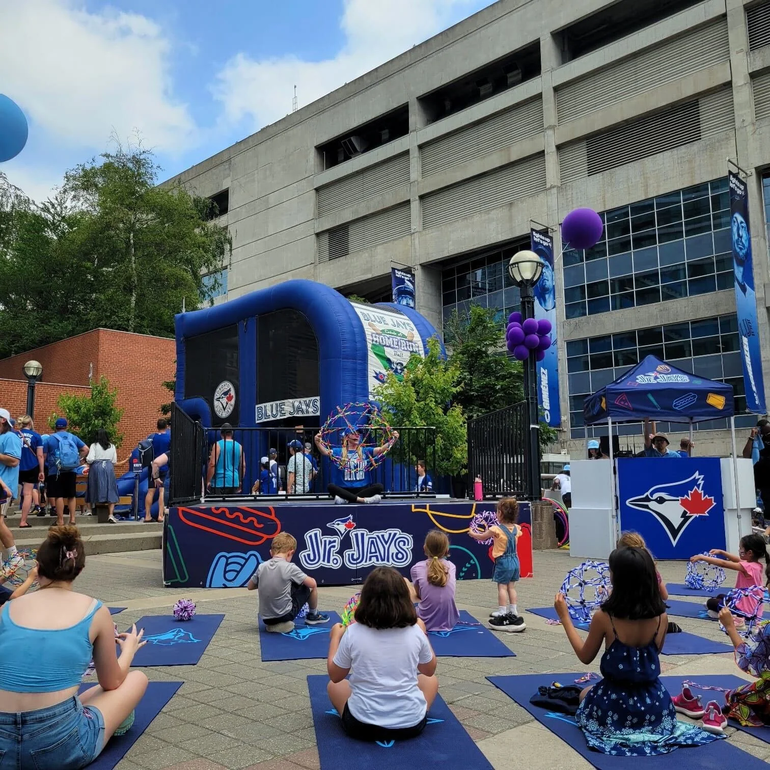 Children participating in an outdoor yoga session at a Toronto Blue Jays event, seated on blue mats with adults and a stage with Blue Jays branding in the background.