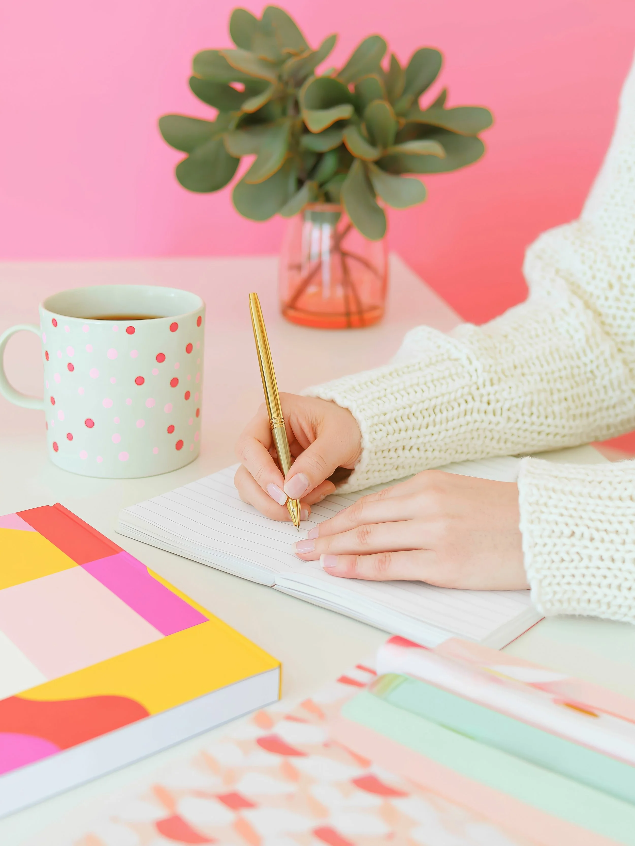 A person holding a spiral-bound manual titled "Little Yogi's Manual & Curriculum" with colorful dots on the cover, sitting on a light wooden table with a glass of water, glasses, and a pen nearby.