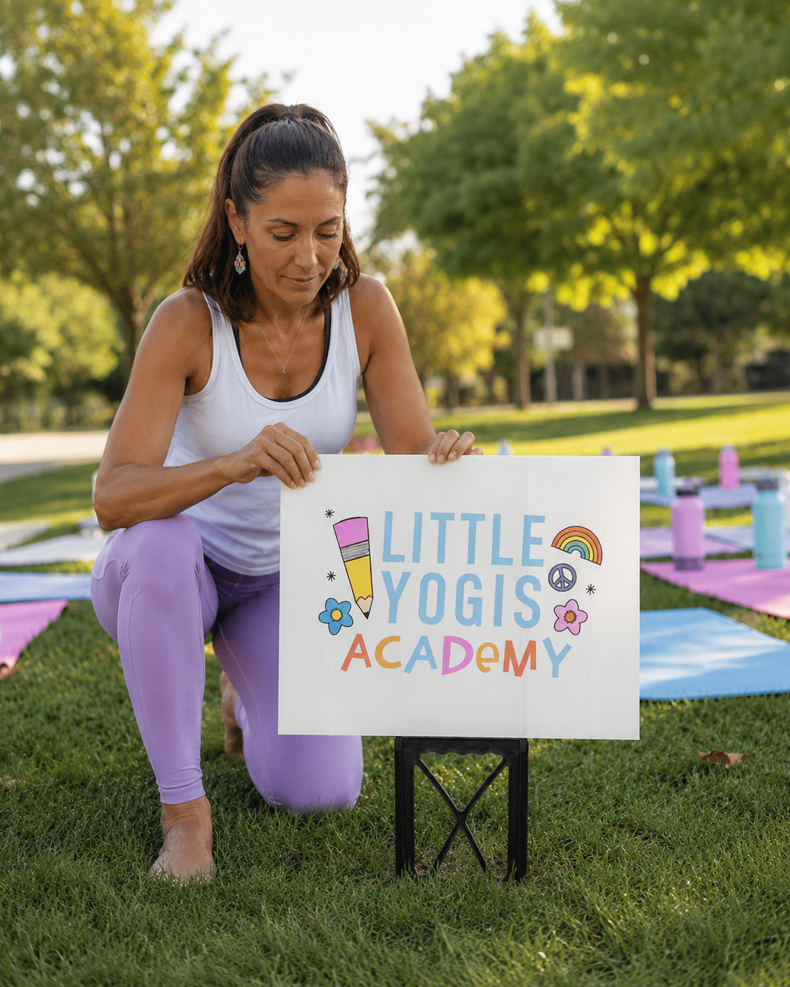 A woman kneeling on grass outdoors holding a colorful sign that says 'Little Yogis Academy' with illustrations of a pencil, rainbow, peace sign, and flowers. There are yoga mats and water bottles set up behind her.