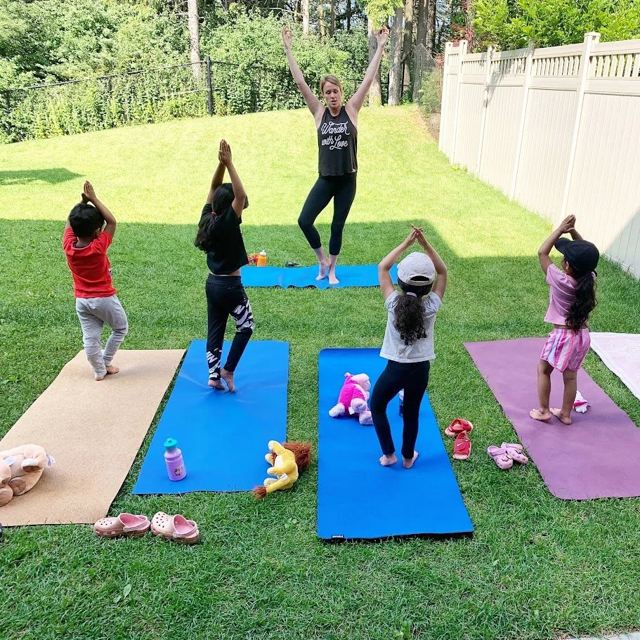 A group of children practicing yoga outdoors on colorful mats with an instructor standing on a yoga mat. The children and instructor are doing a pose with their hands raised above their heads, in a backyard surrounded by a white fence and green trees