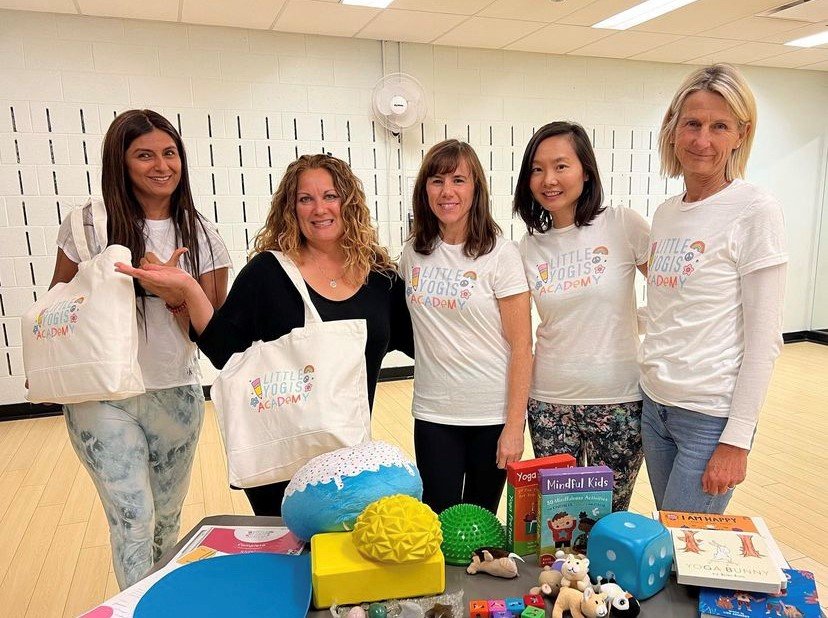 Five women standing indoors behind a table with children's toys and books, some wearing white t-shirts with a logo that says 'Little Yogis Academy', smiling at the camera.