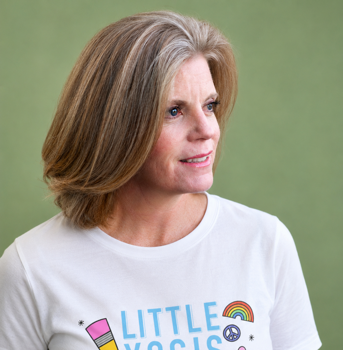 A woman with shoulder-length light brown hair and blue eyes wearing a white T-shirt with colorful rainbow and peace symbol designs on a green background.
